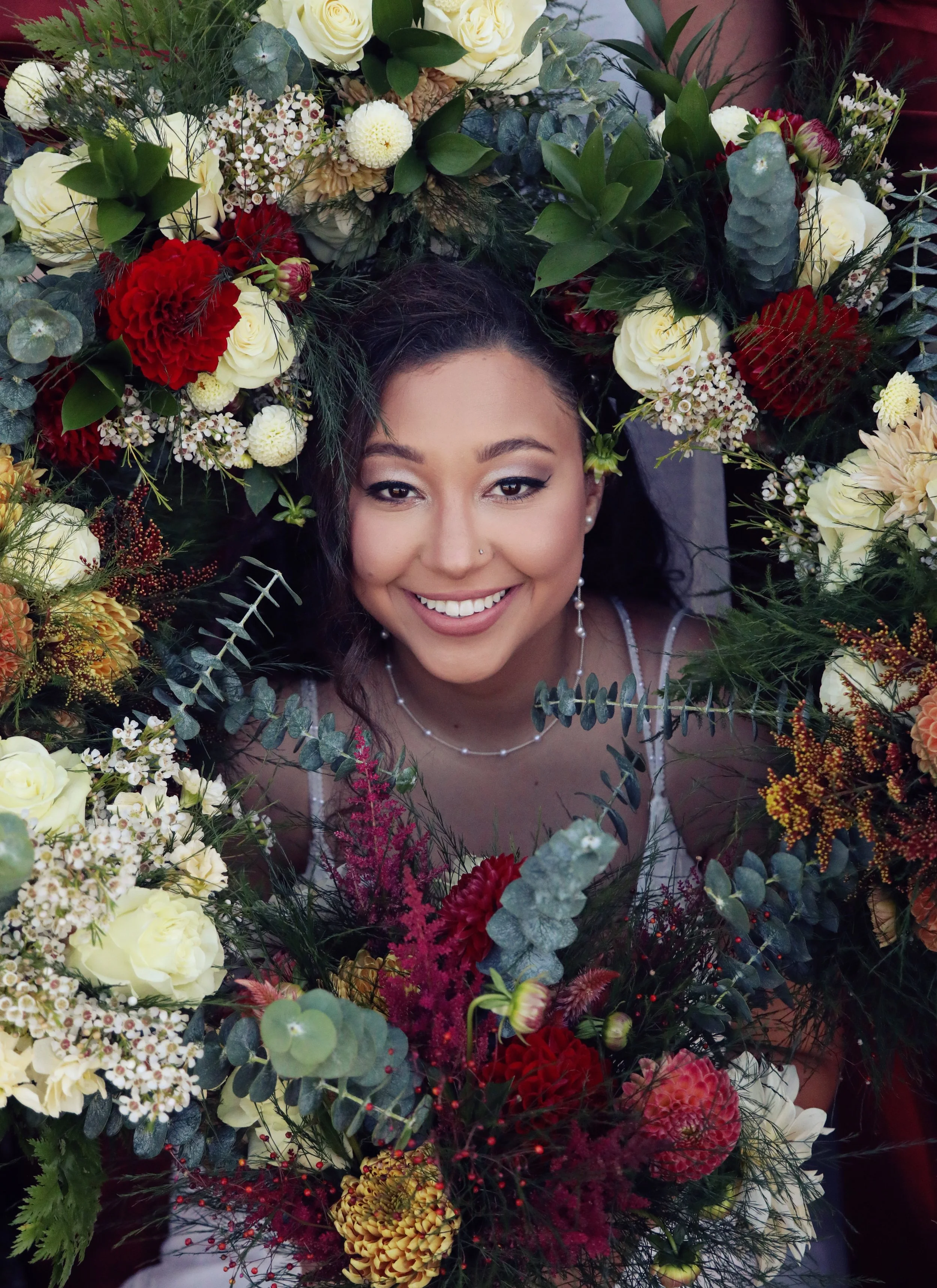 A woman smiling and wearing jewelry surrounded by a large colorful flower arrangement.