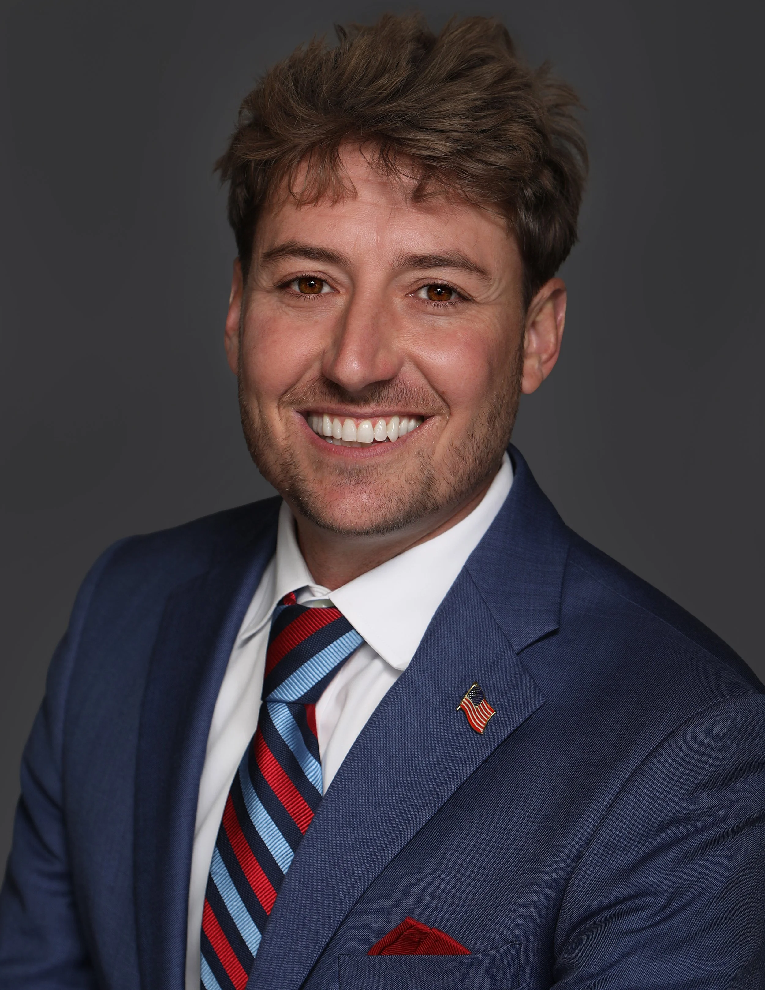 Portrait of a man with light brown hair and a beard, smiling, wearing a blue suit, white shirt, striped red, blue, and black tie, and a small American flag pin on his lapel against a gray background.