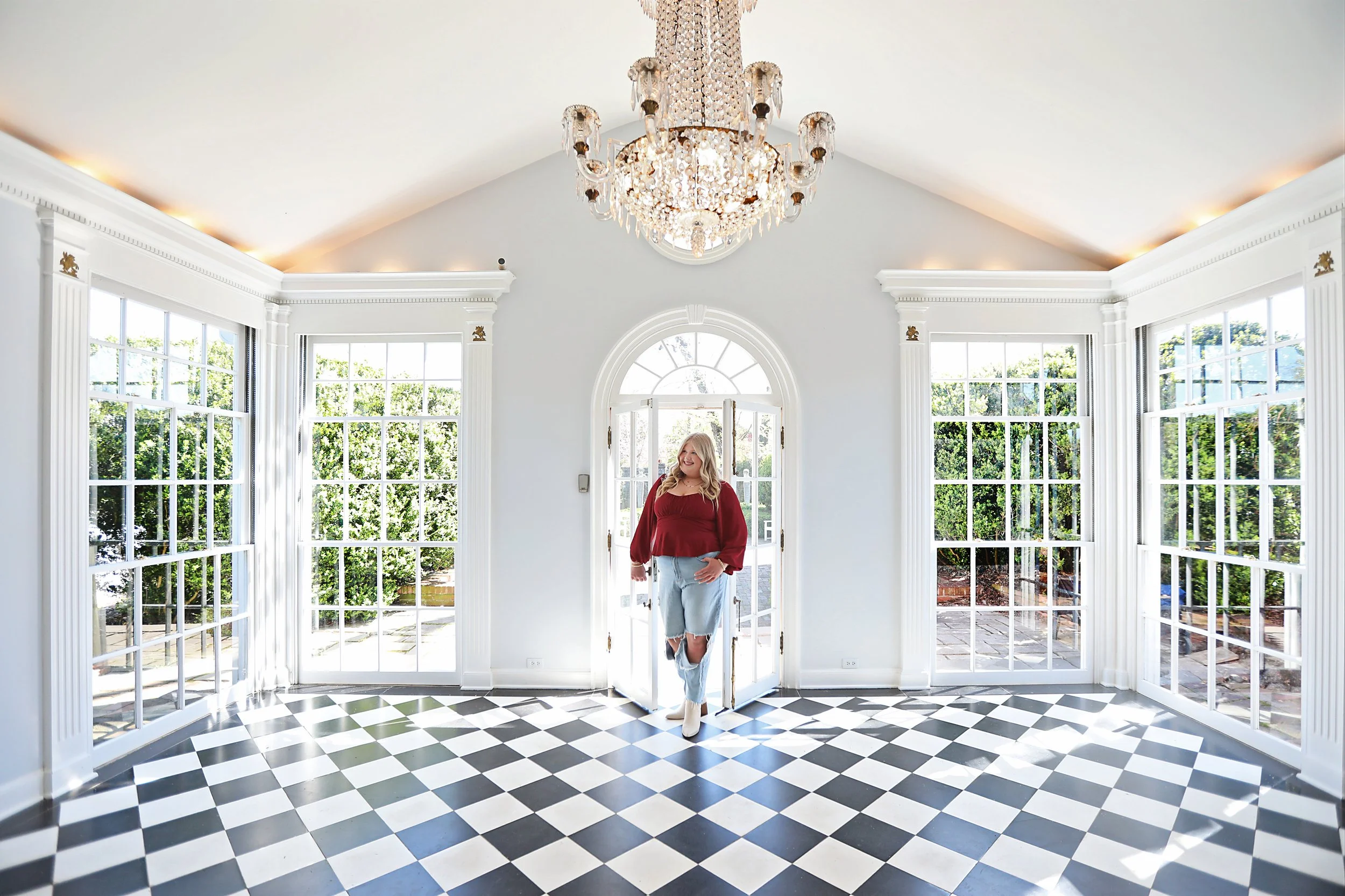A woman with blonde hair wearing a red top, ripped jeans, and beige boots standing in the doorway of a bright, spacious room with large windows and a black-and-white checkered floor, sunlight streaming in.