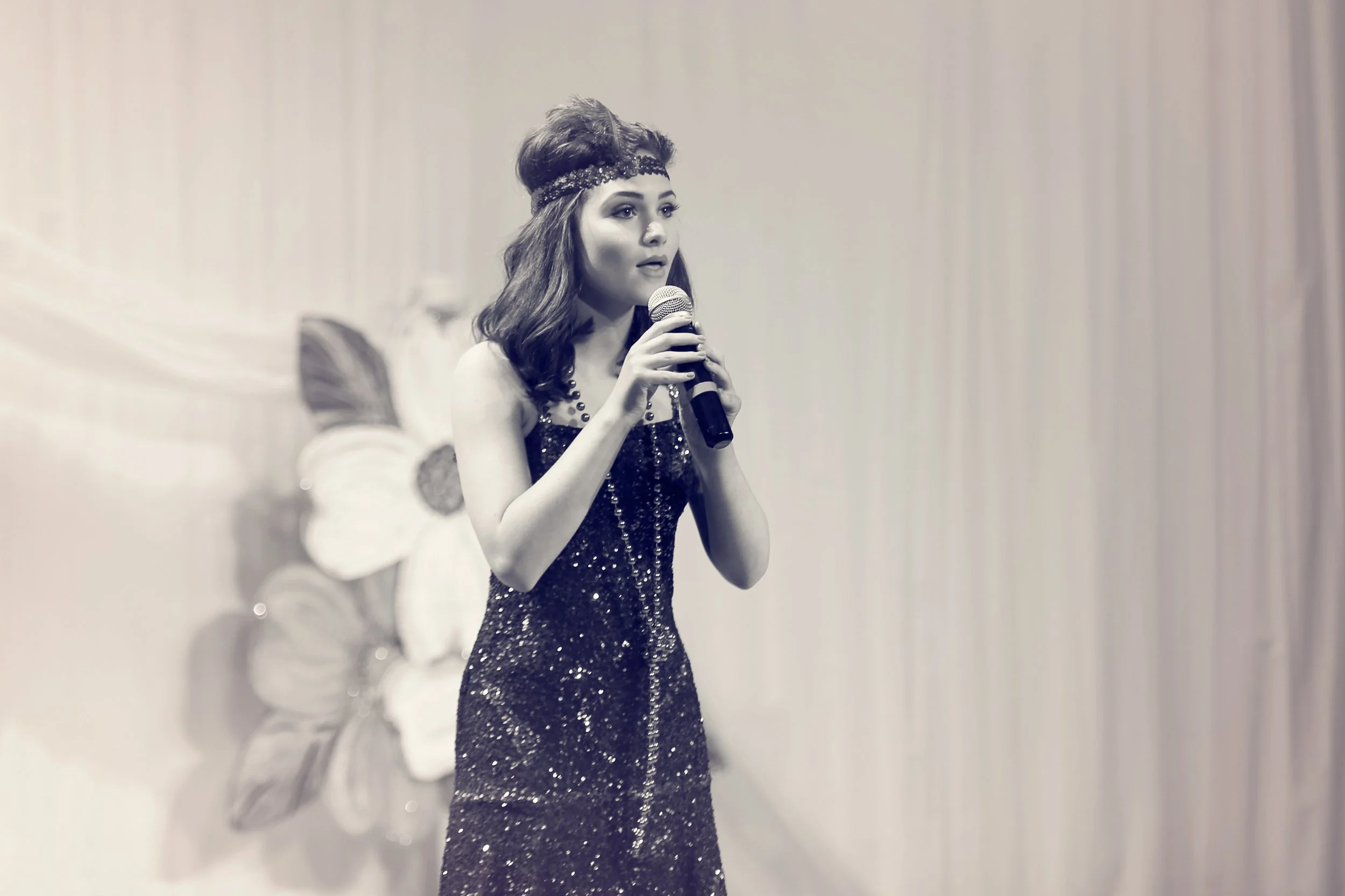Young woman singing into a microphone on stage, wearing a sparkly dress and a decorative headband, with a floral background.