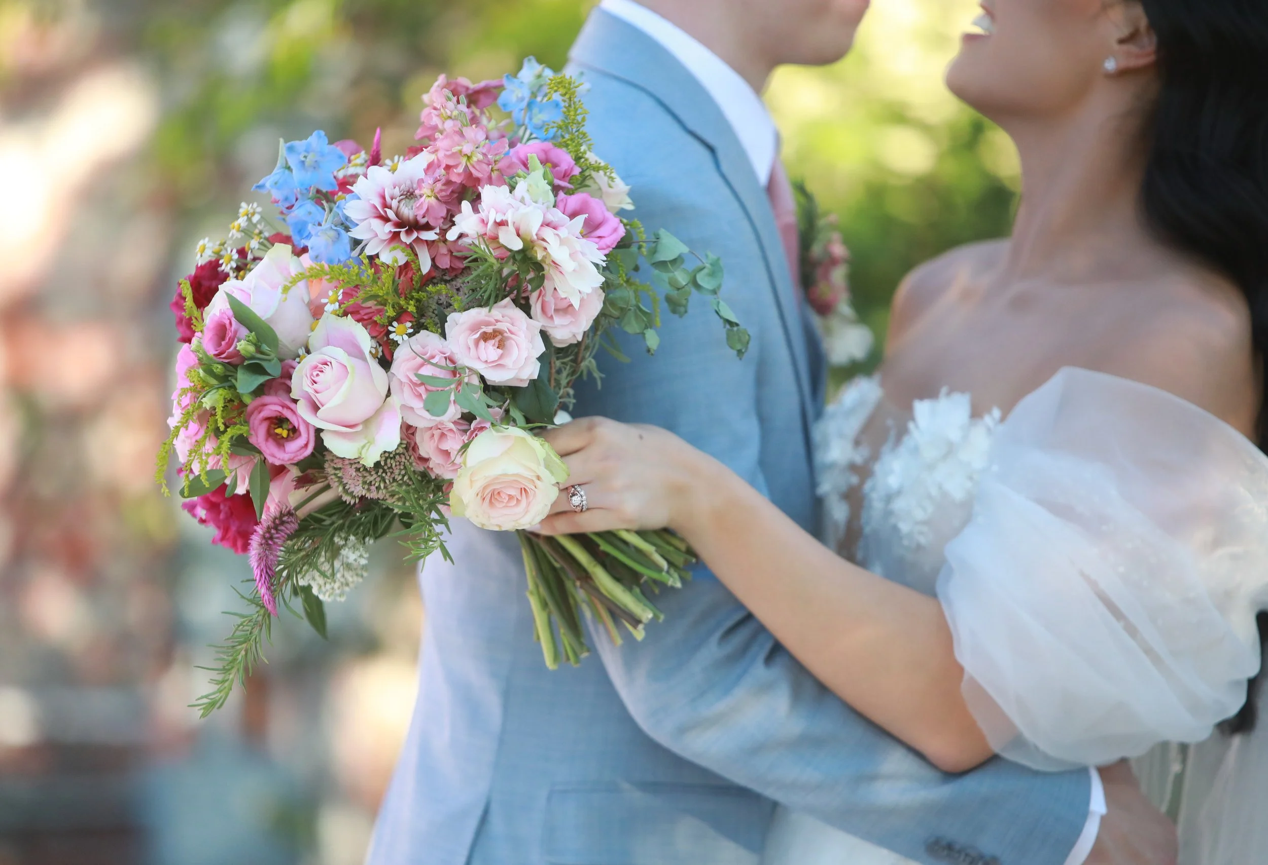 A bride and groom at their wedding, the bride holding a bouquet of pink, white, and blue flowers, and wearing a white wedding dress with lace and tulle details.