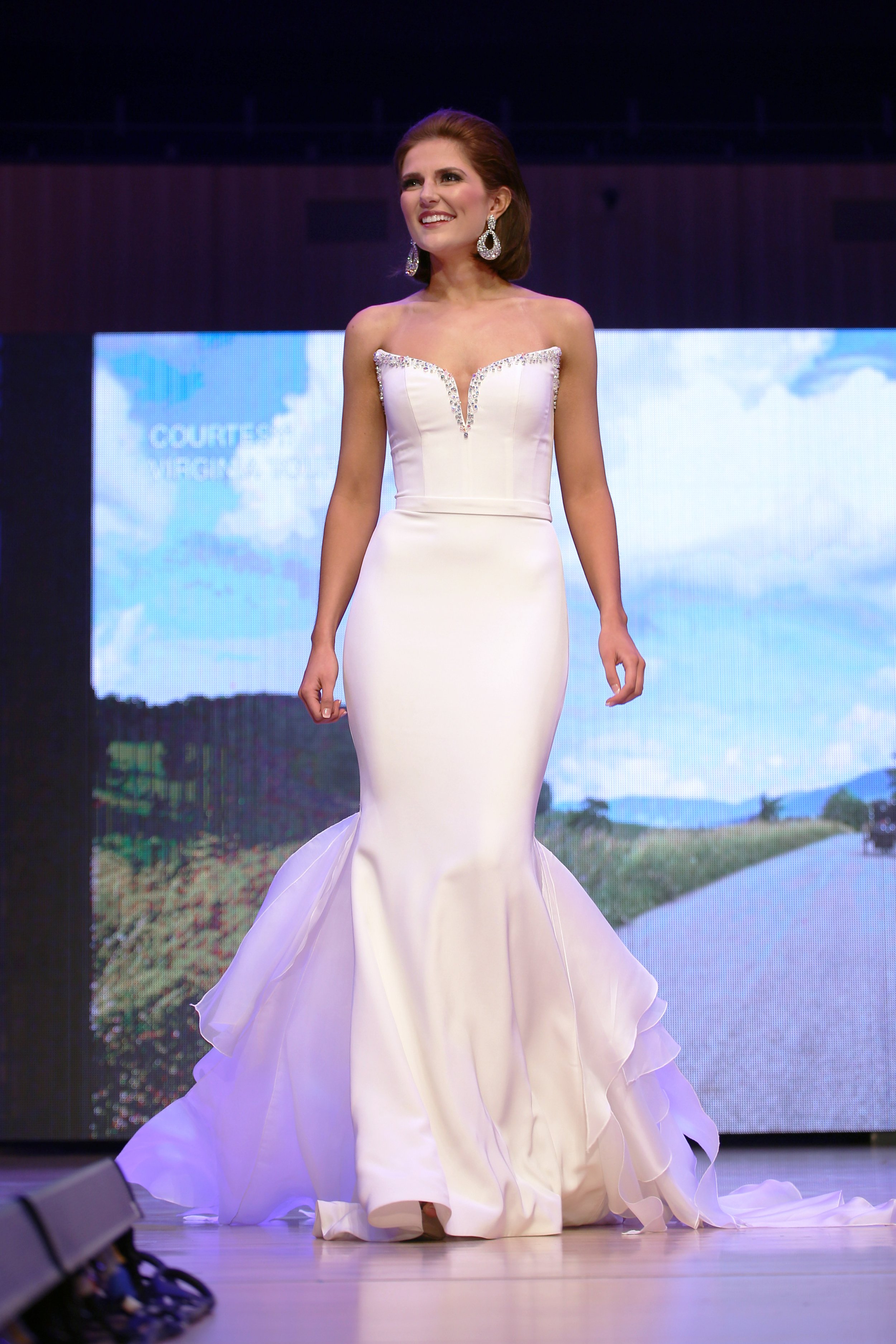 A woman in a white, strapless wedding gown with a mermaid silhouette and ruffled hem, walking on a runway stage with a digital screen in the background showing a rural landscape and blue sky.