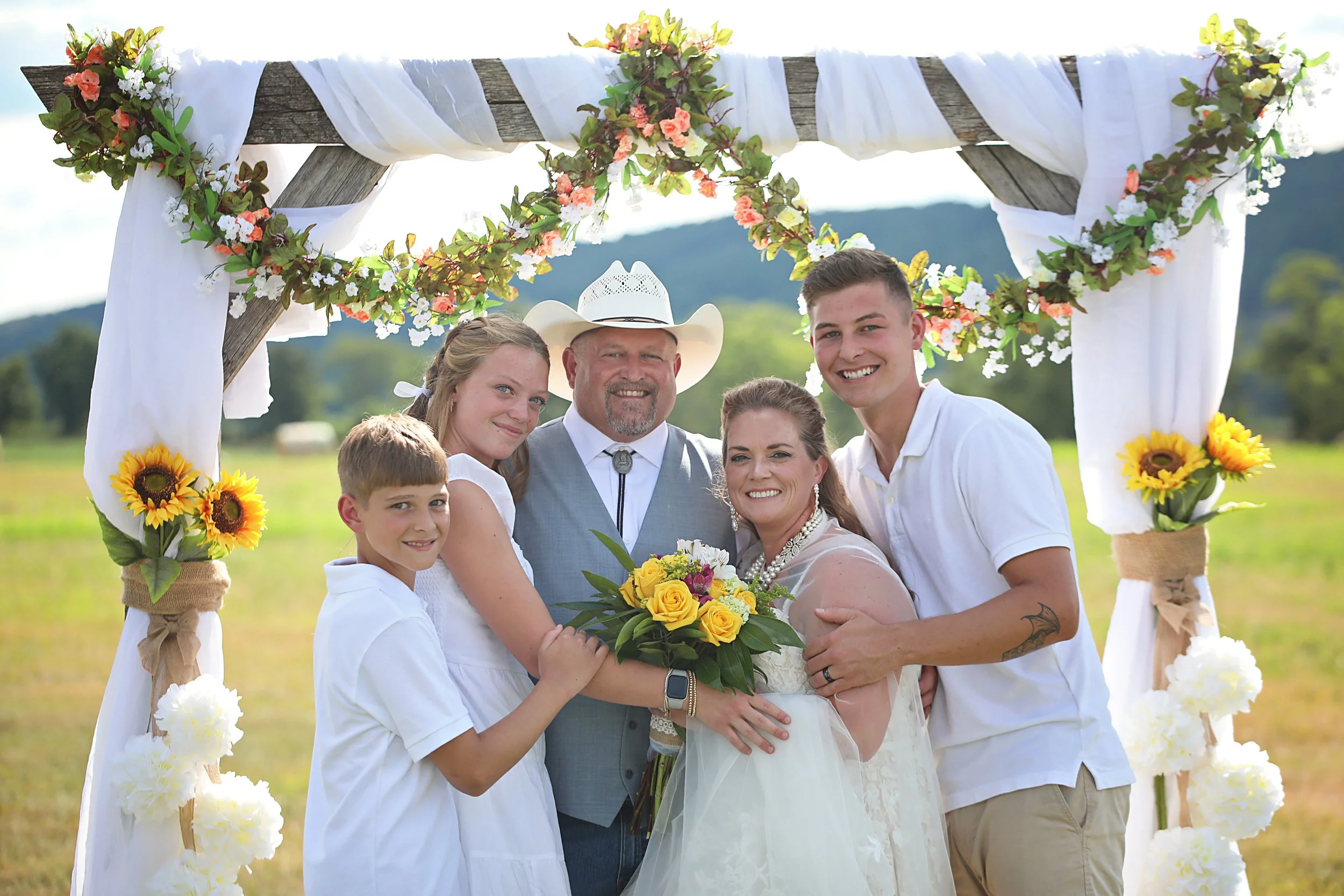 A family posing in a field during a wedding ceremony with a decorated wooden arch, white fabric, sunflowers, and flowers