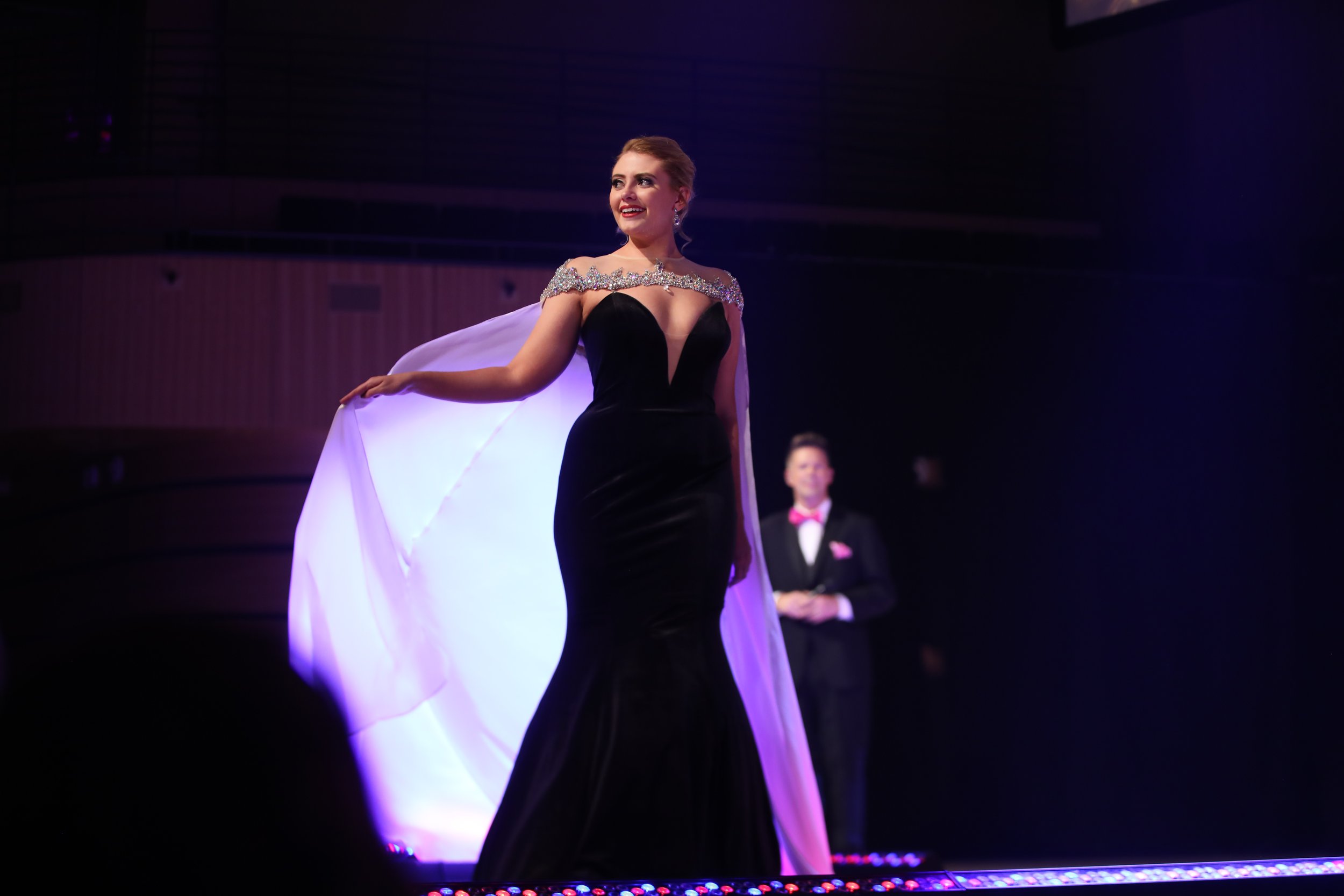 Woman in a black evening gown with sparkly embellishments on the shoulders, holding a white cape, standing on a stage with a man in a tuxedo in the background.