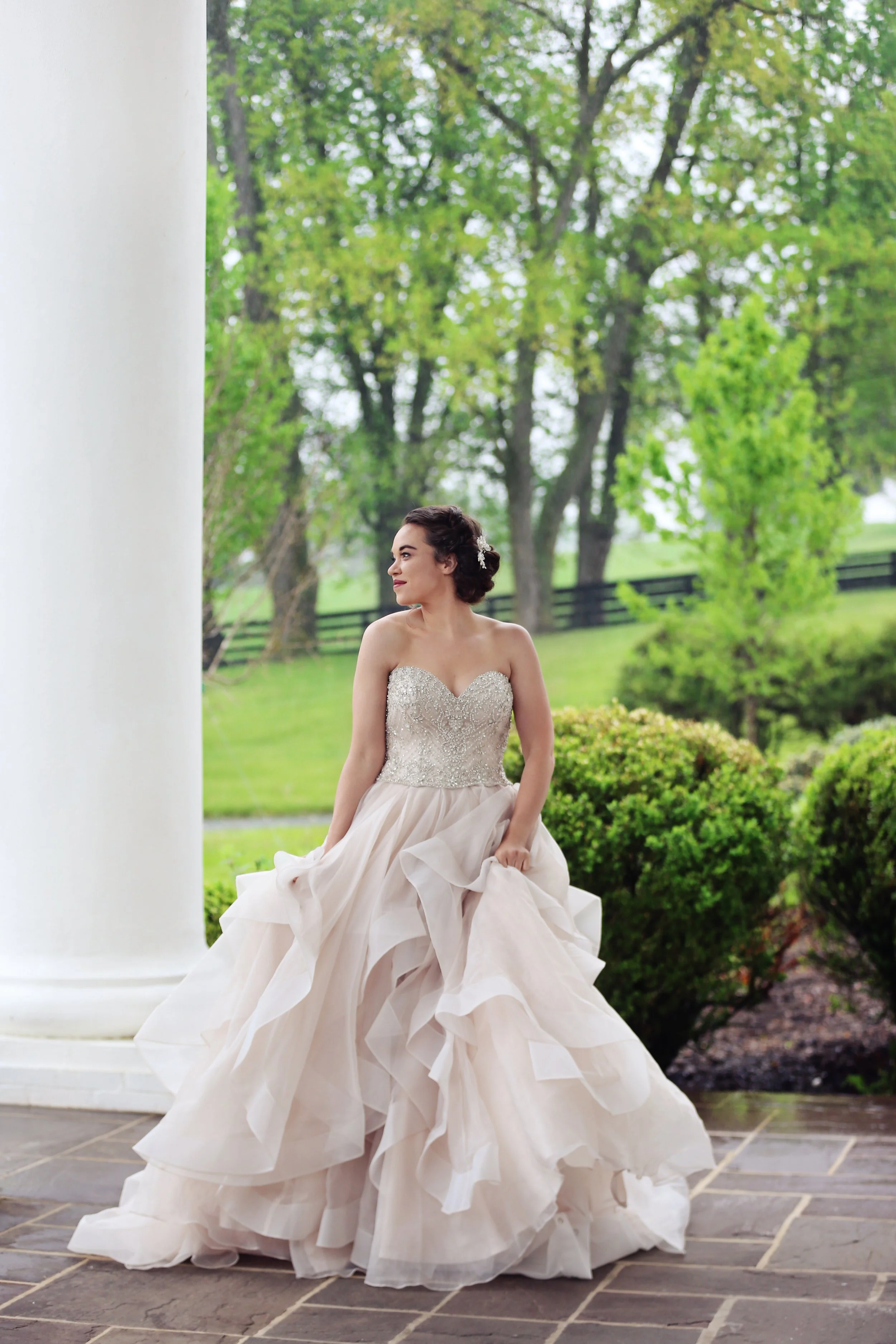 A woman in a wedding dress sitting outdoors near a white pillar, with trees and green bushes in the background.