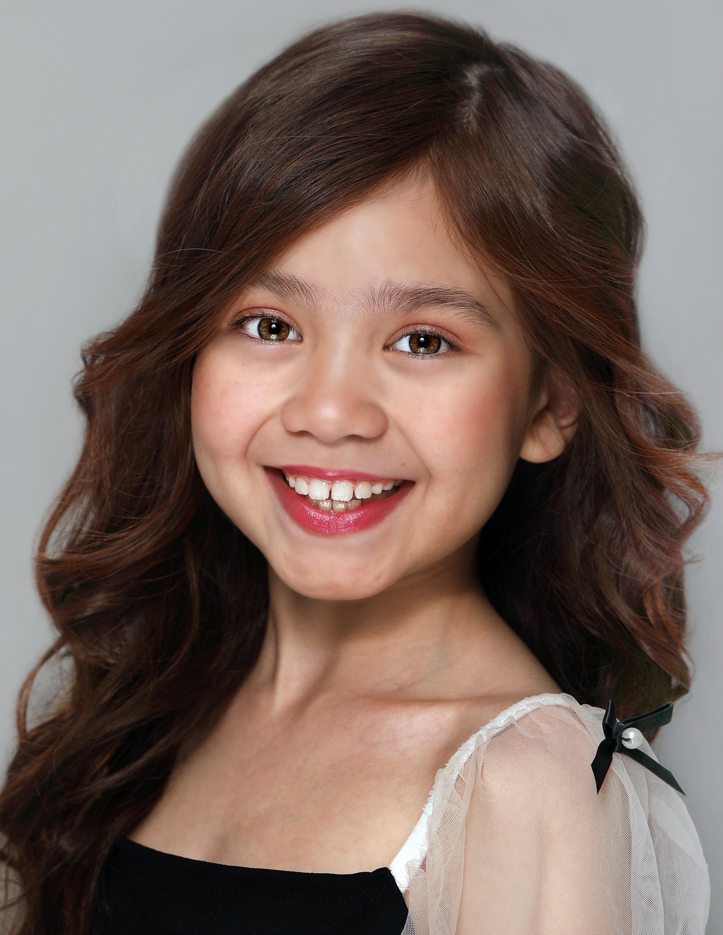 A young girl with long, wavy brown hair smiling at the camera, wearing a black top with a sheer, cream-colored sleeve accented with a small black bow.