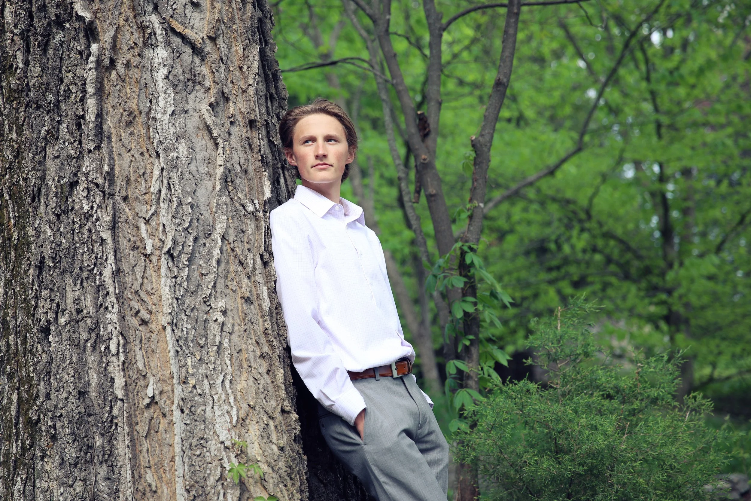 A young man in a white dress shirt and gray pants leaning against a large tree trunk in a lush green forest.