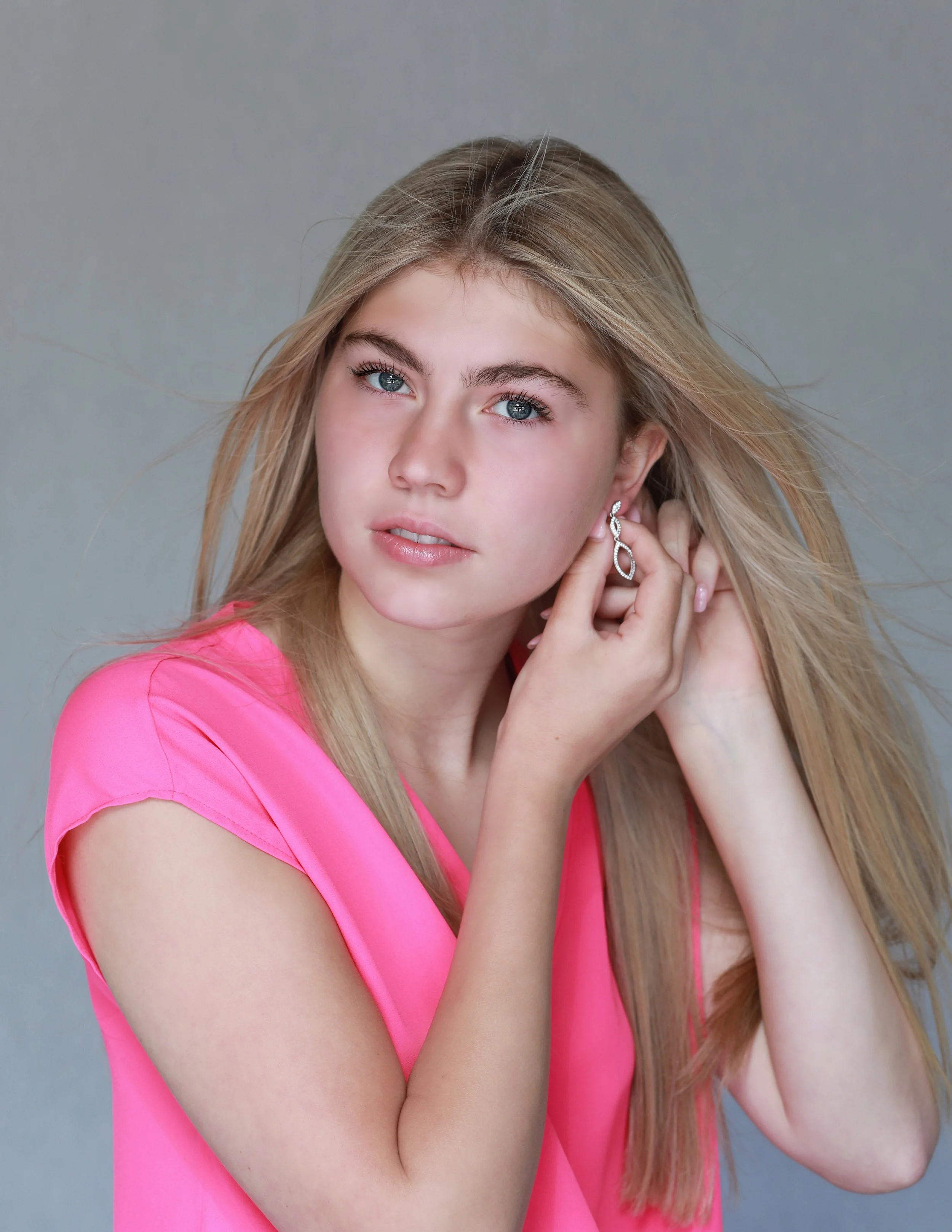 A young woman with long blonde hair and blue eyes, wearing a pink top, adjusting an earring.