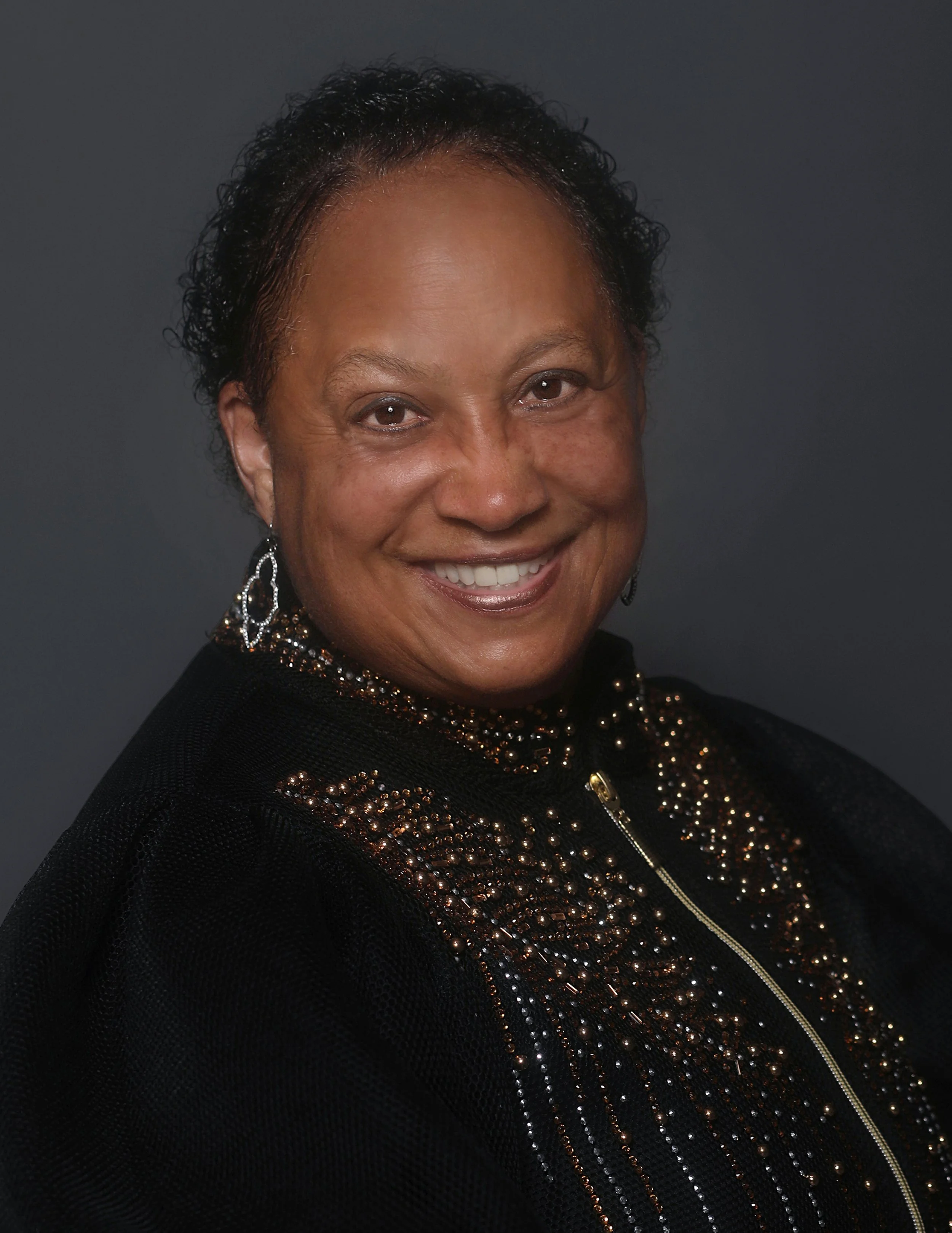 A portrait of an smiling African American woman with short curly hair, wearing a black top with embellished gold and black beads and earrings.