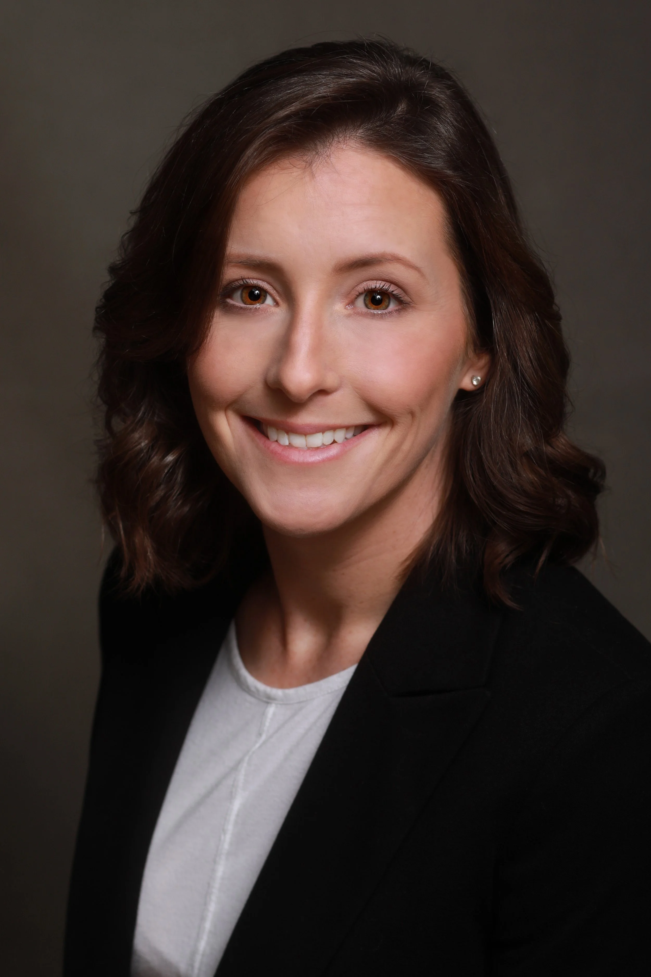 Headshot of a woman with wavy brown hair, wearing a black blazer and white top, smiling against a dark background.
