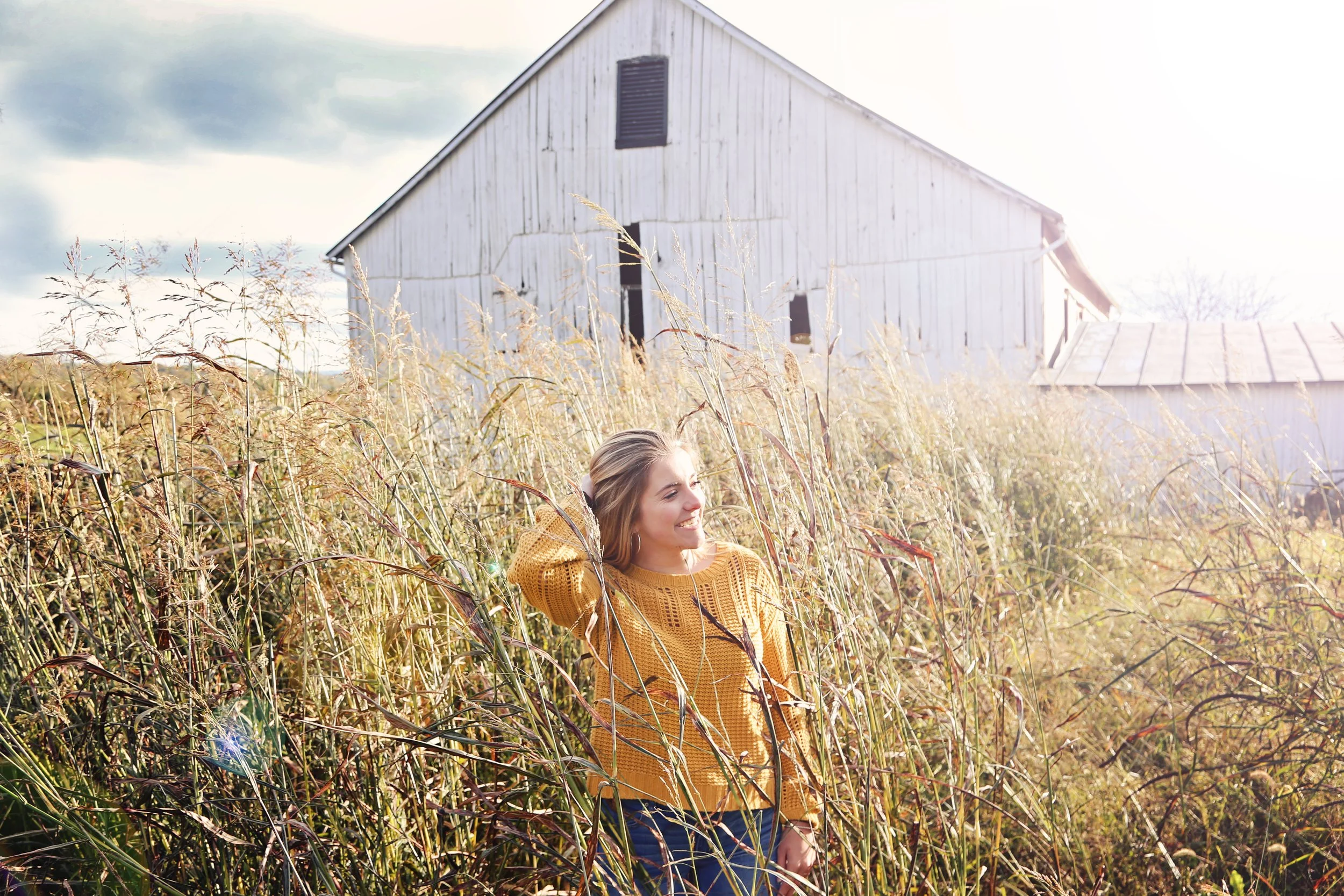 A woman in a mustard yellow sweater standing in tall grass beside a weathered white barn on a sunny day.