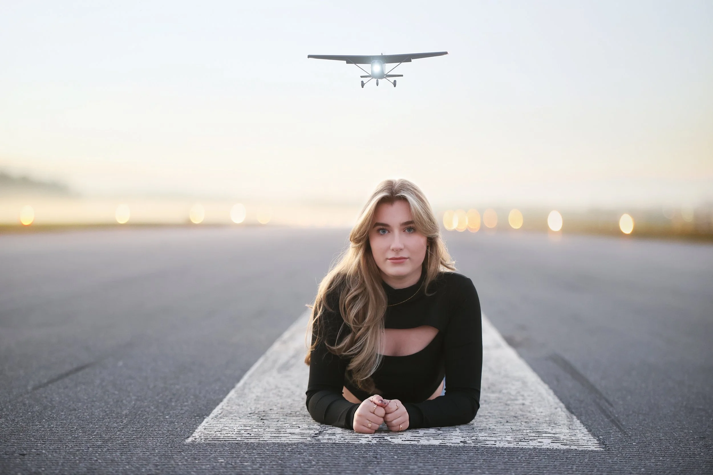 A young woman lying on a runway with a small airplane flying overhead at sunset.