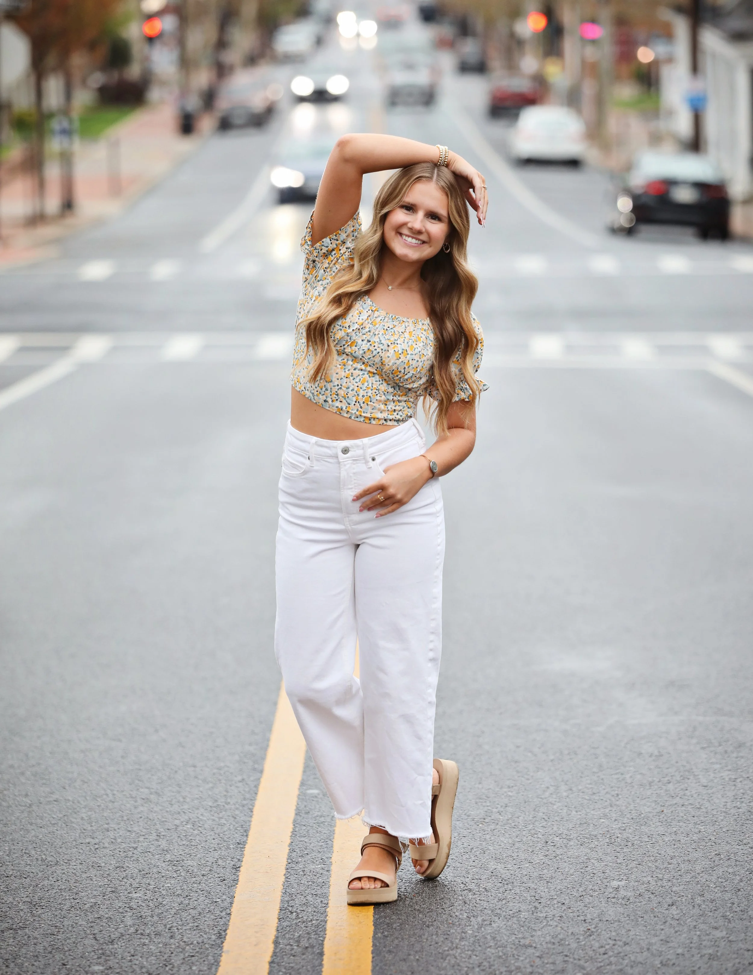 Young woman with long wavy hair posing in the middle of an empty city street, wearing a floral crop top and white jeans, smiling at the camera.