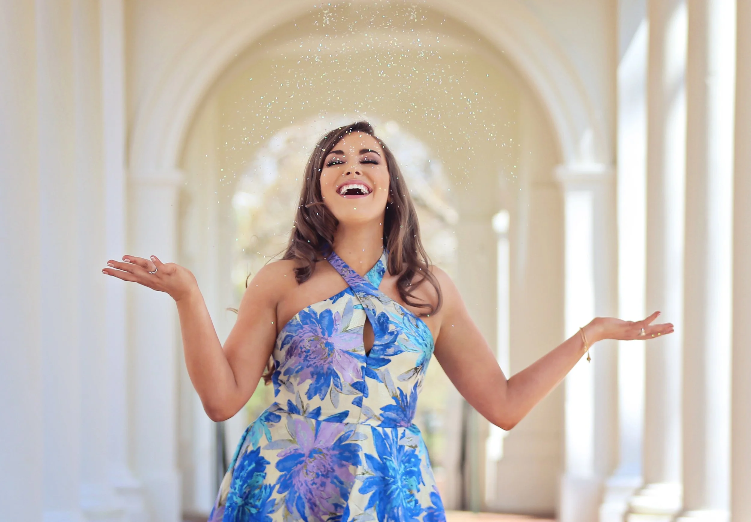 A woman in a floral dress is smiling and throwing confetti in an elegant indoor setting with arches and columns.