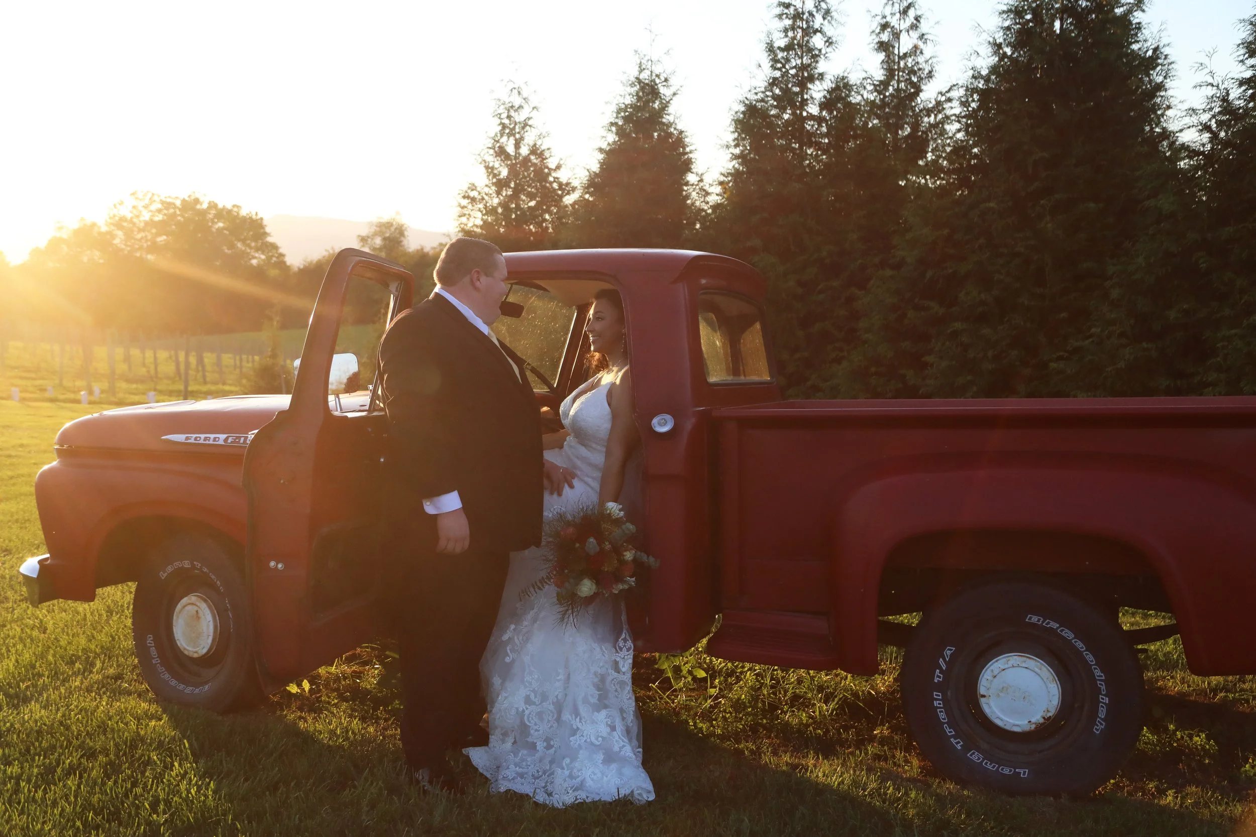 A bride and groom stand beside a red vintage pickup truck in a grassy field at sunset, with trees in the background.