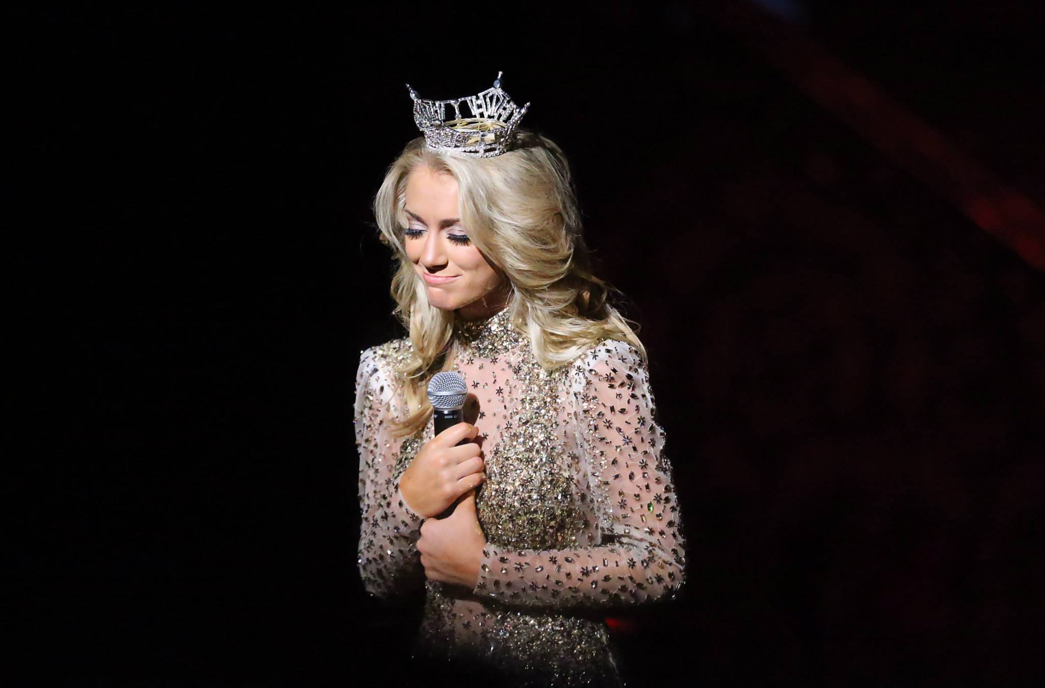 A woman with long, wavy blonde hair, wearing a sparkly, ornate dress and a tiara that says 'Miss Universe', holding a microphone with both hands, standing against a dark background. She appears emotional or reflective.