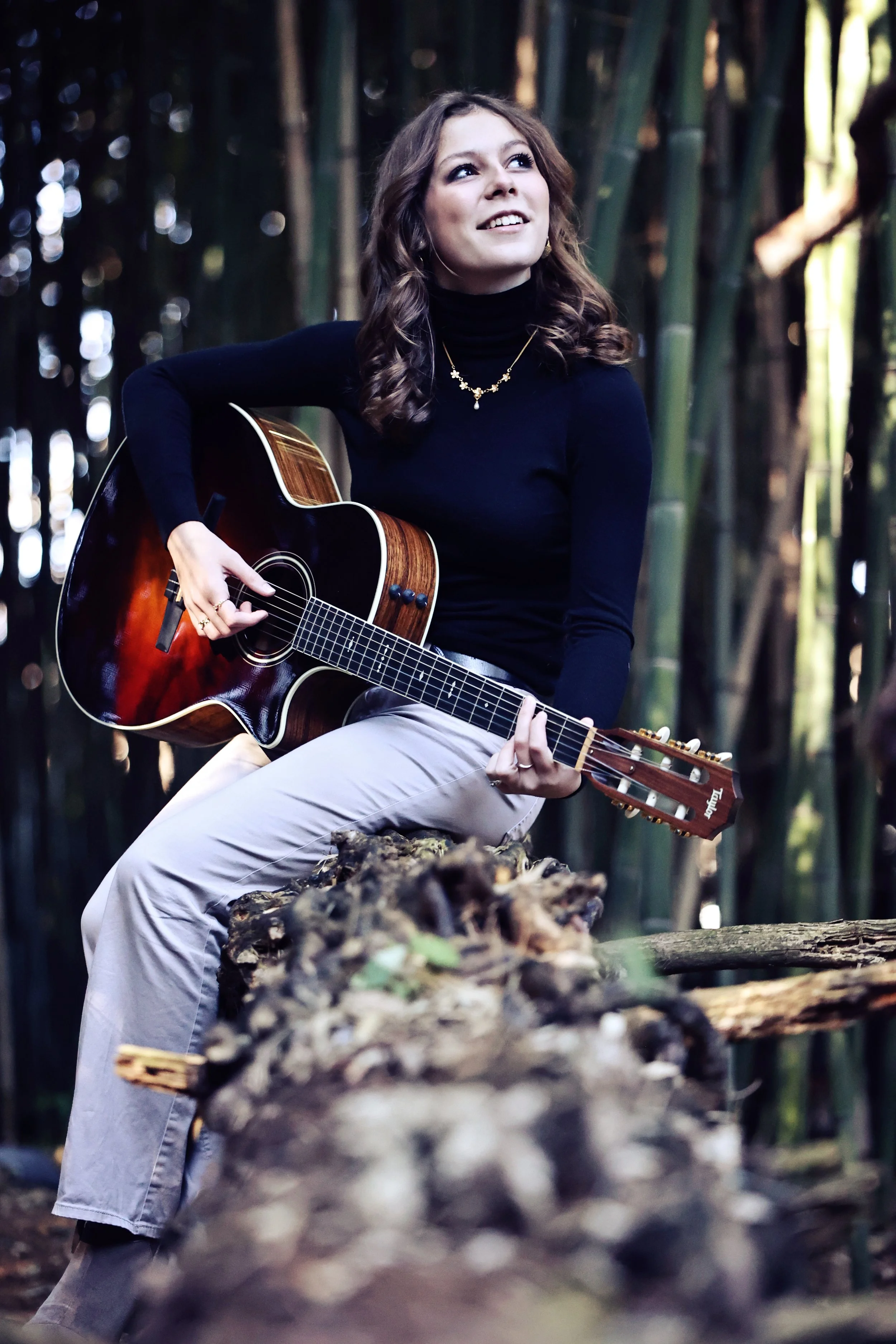 A woman sitting on a log in a forest, playing an acoustic guitar and smiling, with bamboo trees in the background.