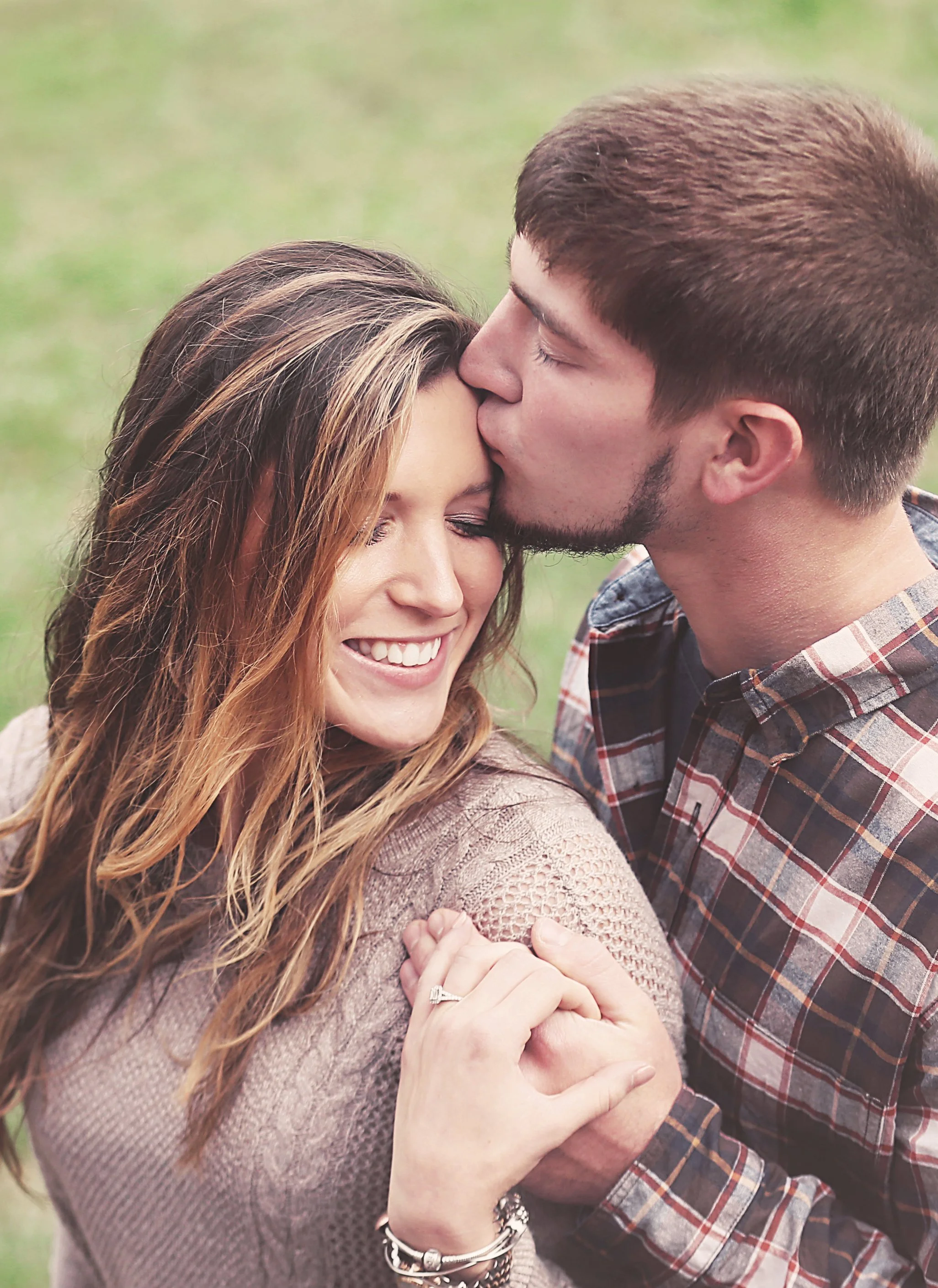 A man kisses a woman's forehead while she smiles with her eyes closed outdoors.