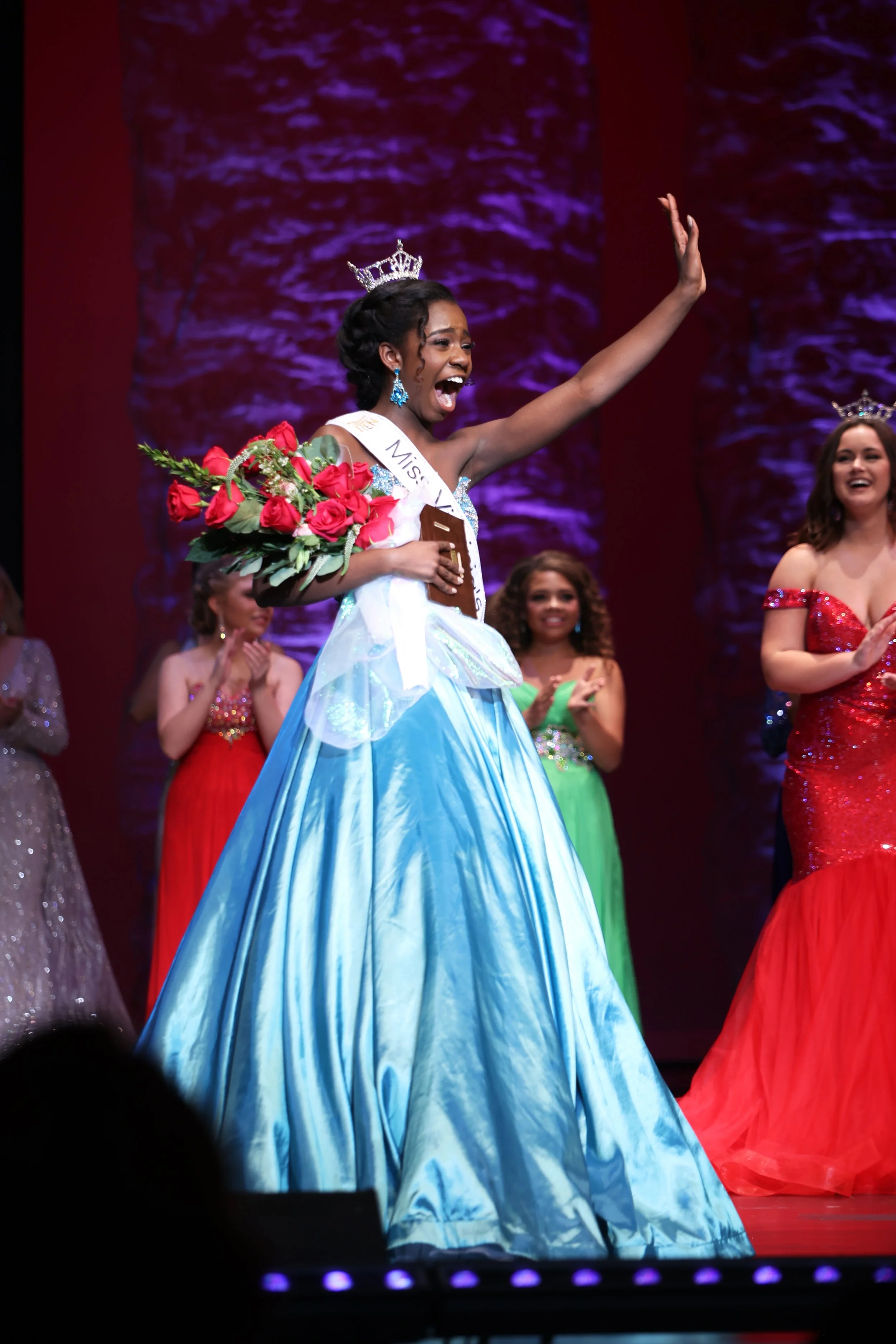 Miss America pageant winner wearing a blue gown, crown, and sash, holding a bouquet of roses, raising her hand in victory, with other contestants in bright gowns applauding on stage.