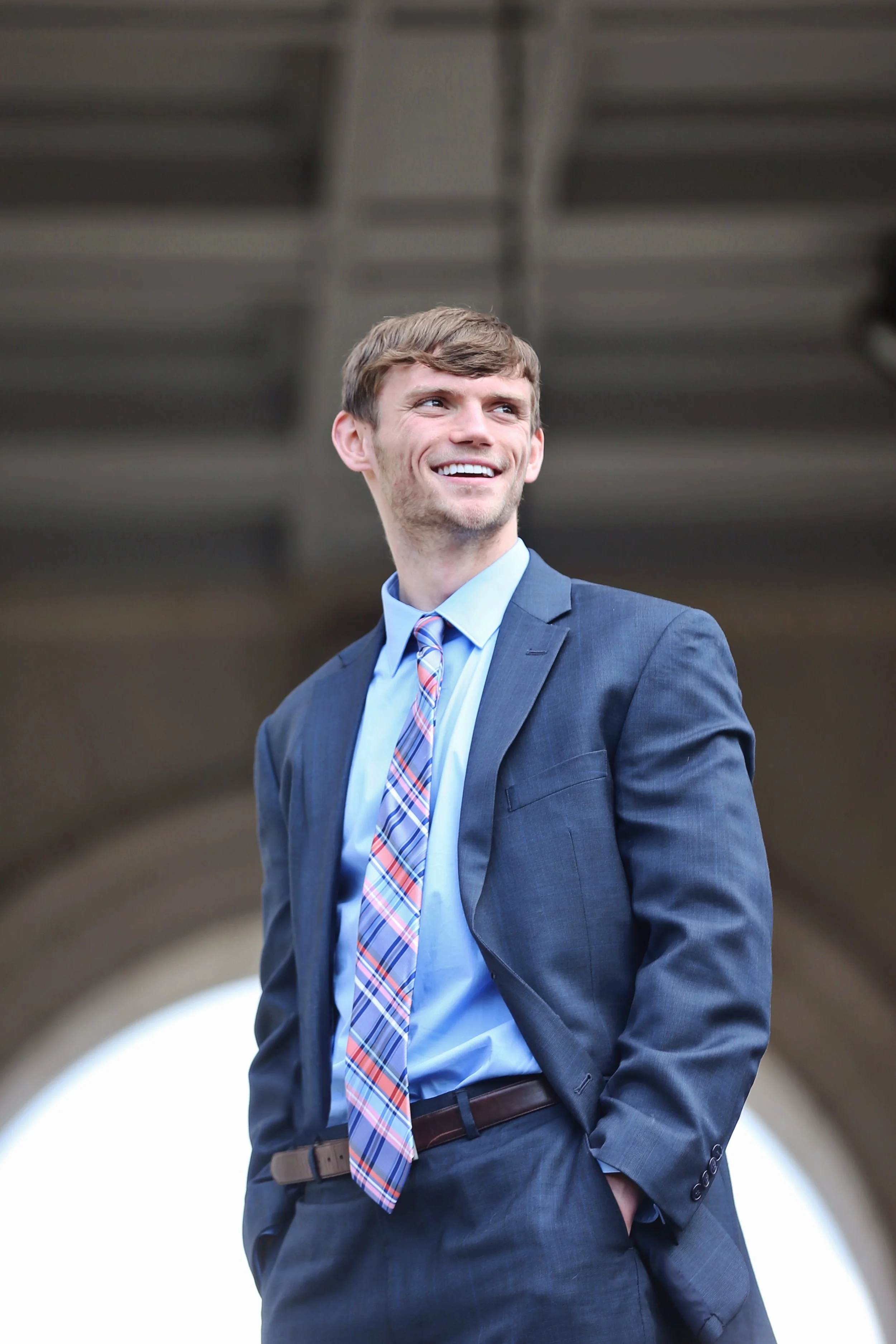 A young man in a suit smiling outdoors under a bridge.