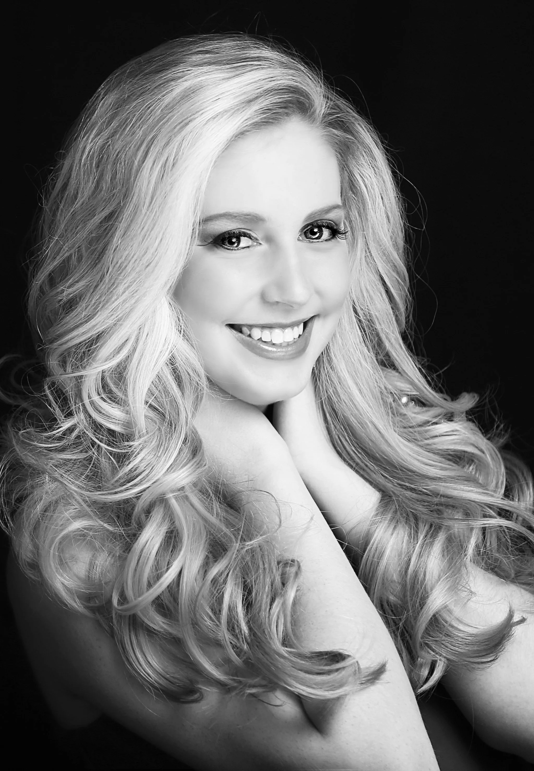 Black and white portrait of a young woman with long, wavy blonde hair, smiling and resting her chin on her hand.