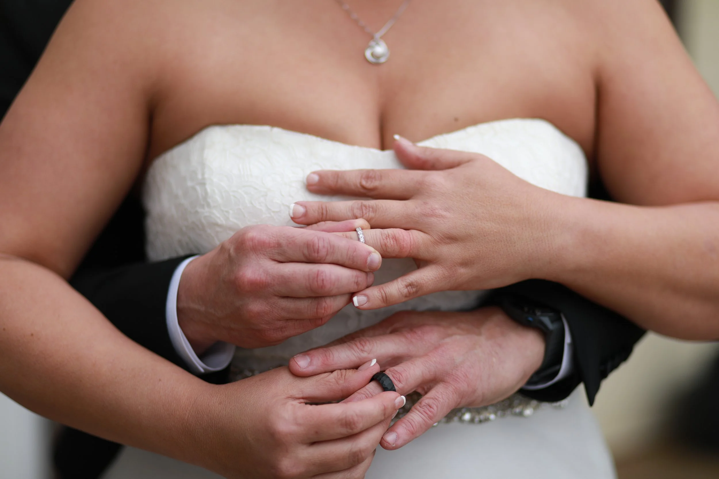 A bride and groom holding hands, showing their wedding rings. The bride is wearing a strapless white wedding dress and a necklace. The groom is dressed in a black suit with a white shirt.