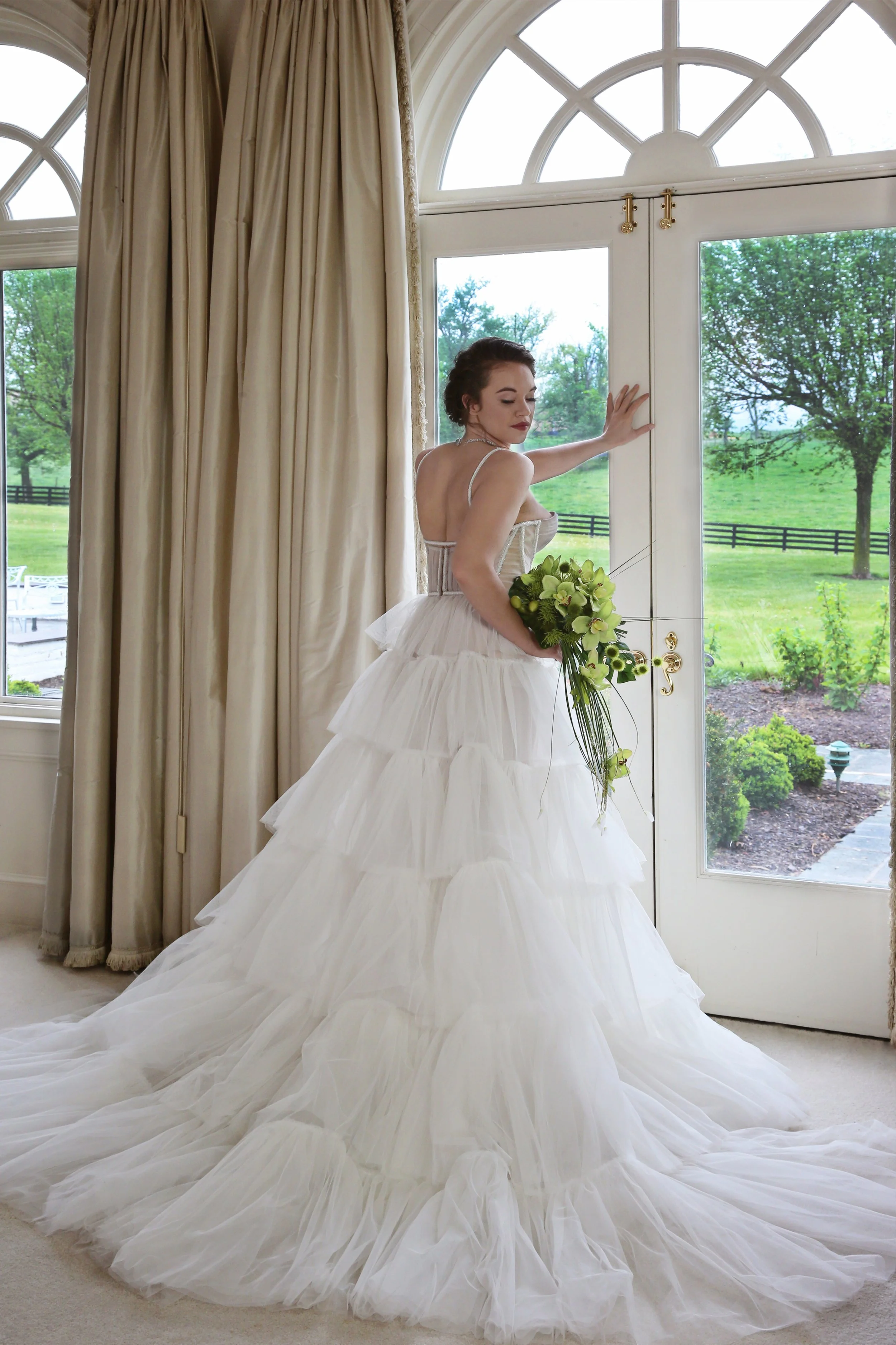 A bride in a white wedding gown holding a bouquet, standing in front of a large window with a view of a green landscape outdoors.