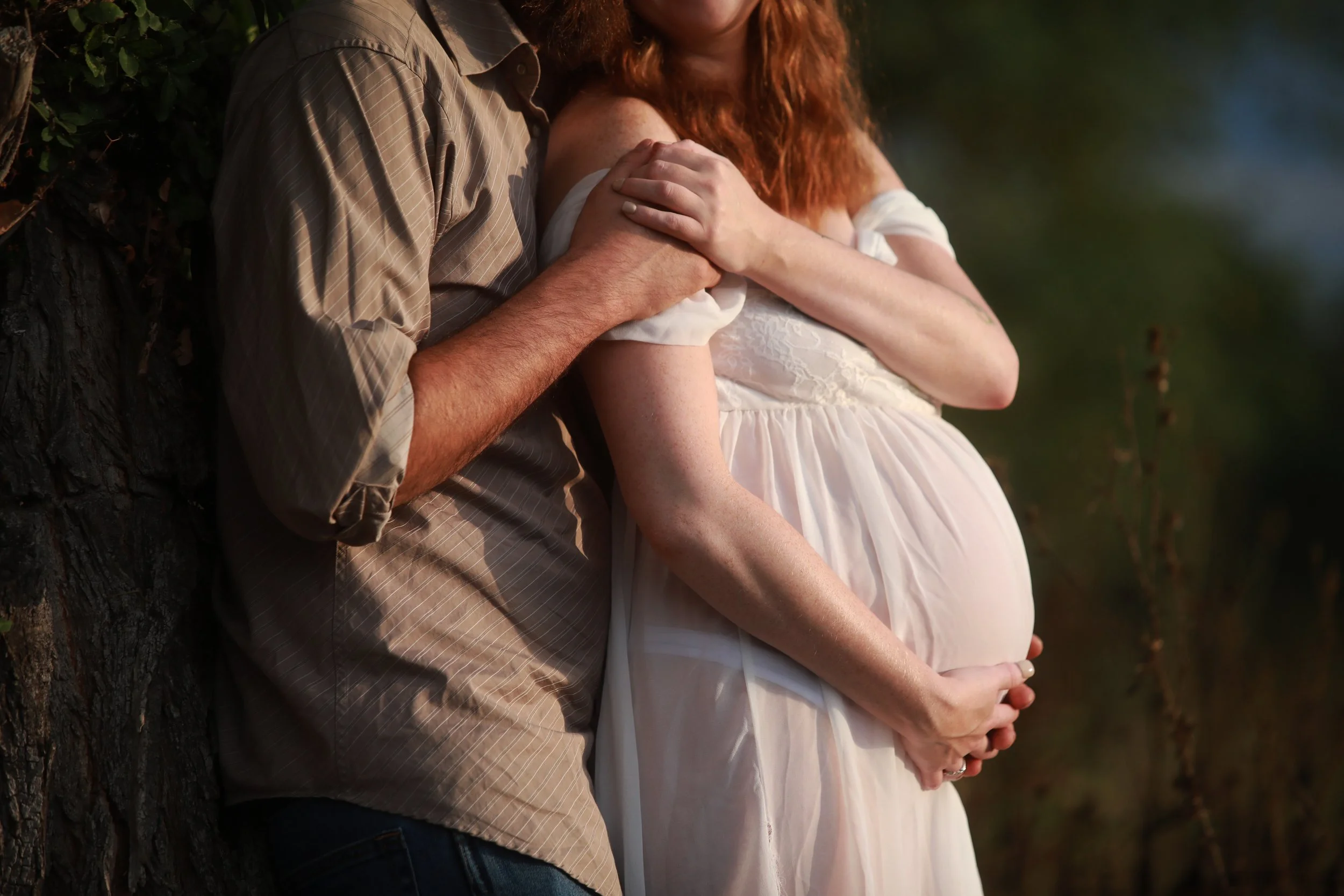 A pregnant woman and a man, possibly her partner, are leaning against a tree outdoors, with the man's hand resting on the woman's shoulder and her hand on her belly.