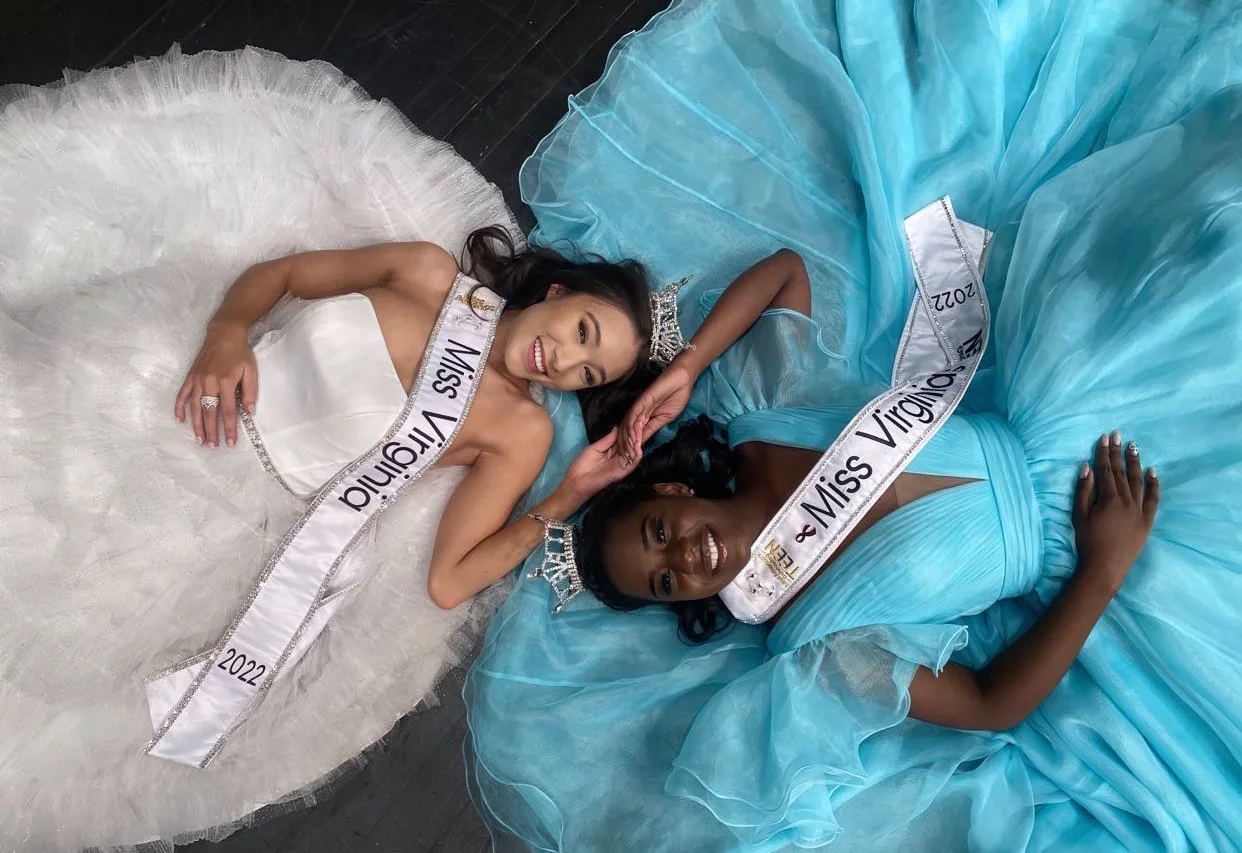 Two women lying on the floor in elegant dresses with 'Miss Virginia 2022' sashes and tiaras, smiling at the camera.