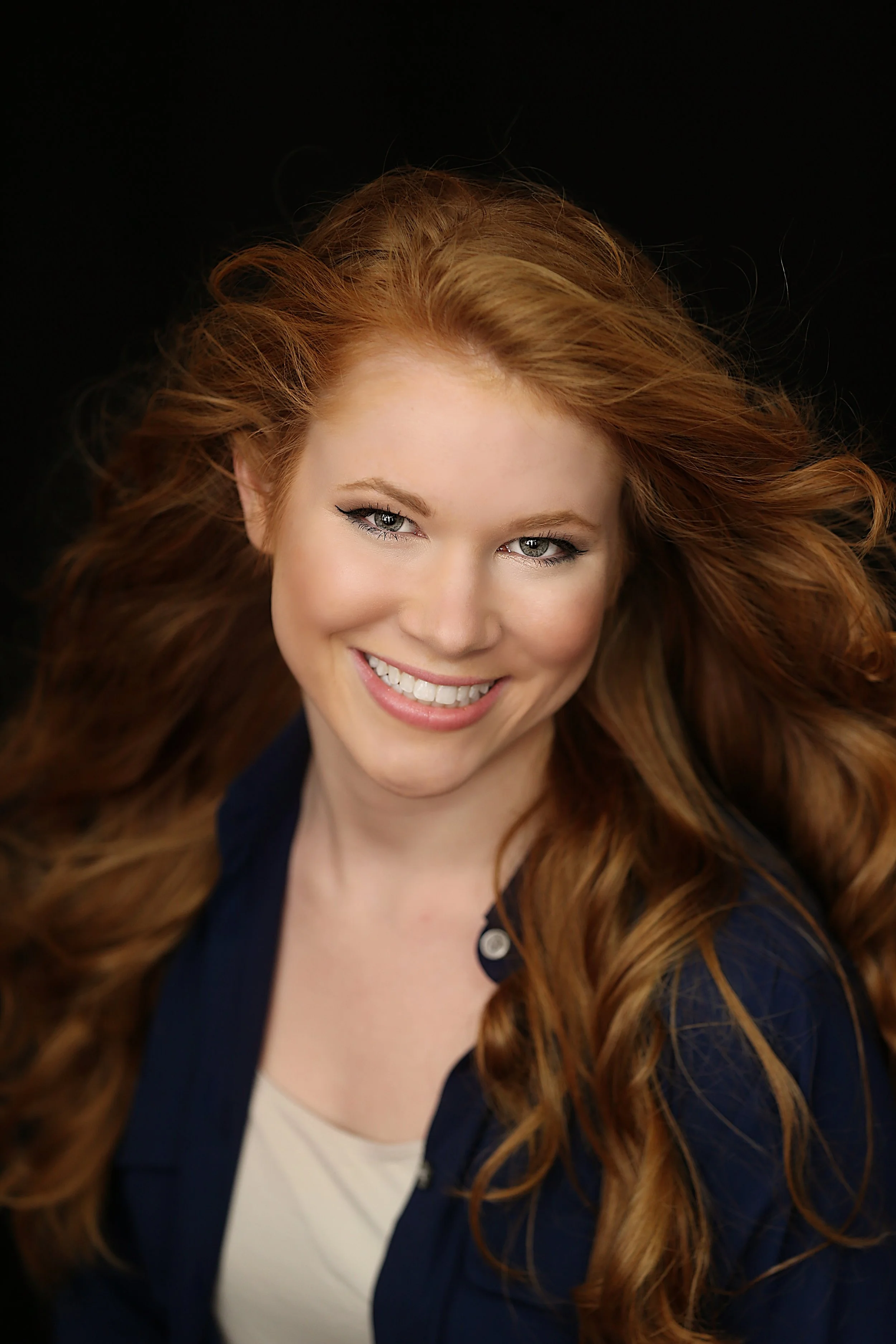 Close-up portrait of a smiling woman with long, wavy red hair, wearing a dark top, against a black background.