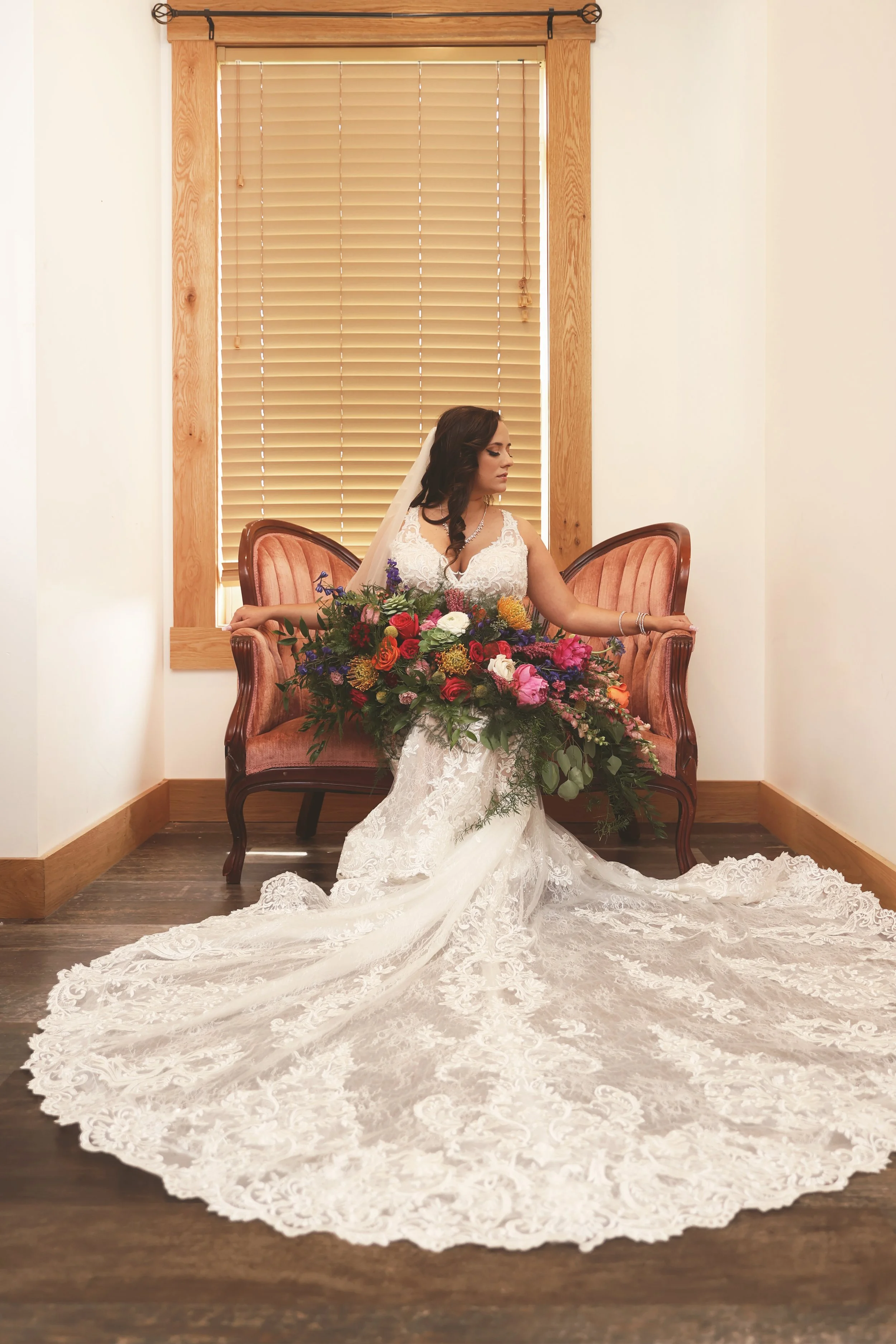 Bride in a lace wedding gown holding a large colorful bouquet, sitting on a vintage pink velvet settee in front of a window with wooden blinds.