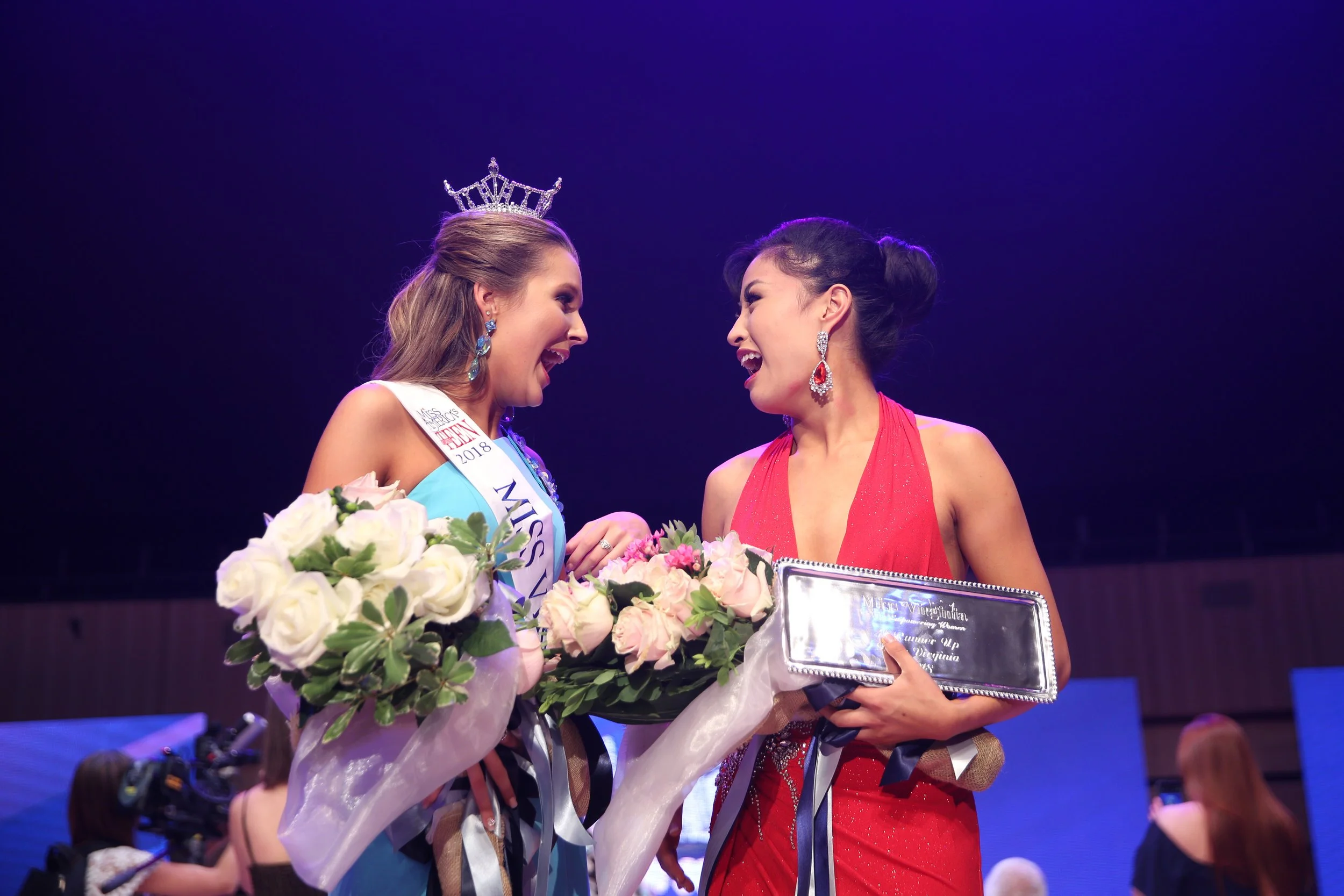 Miss International pageant winner wearing a crown and sash, celebrating with another contestant in a red dress, holding a bouquet of roses and a silver plaque, on stage with a purple backdrop.