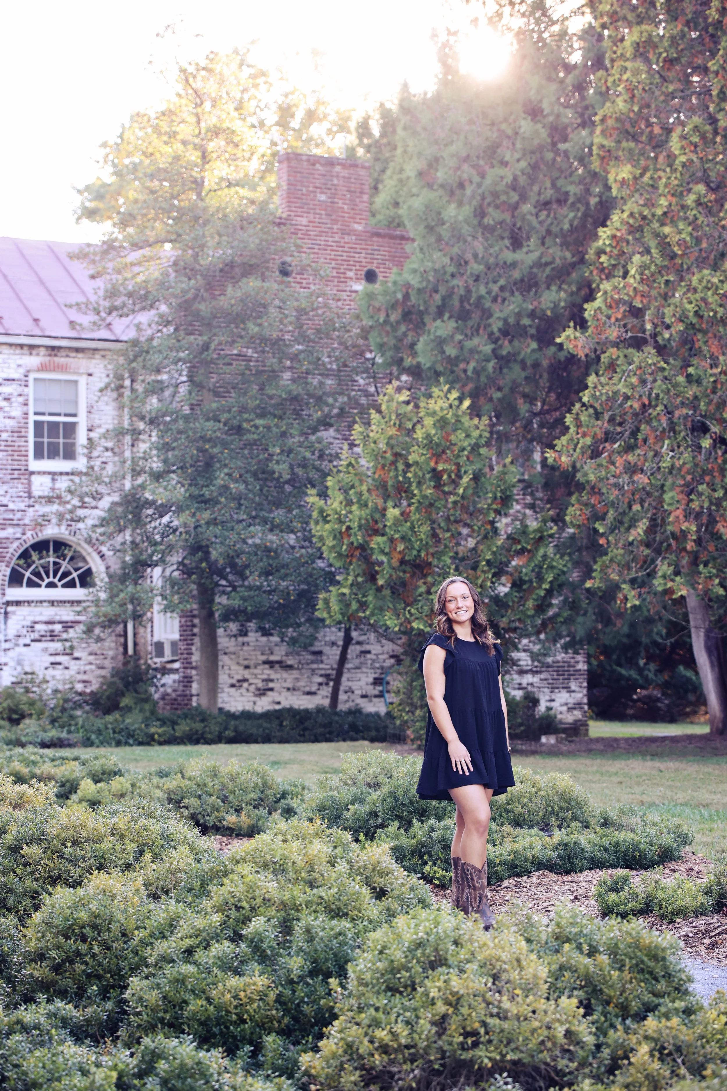A young woman in a black dress and cowboy boots standing in a garden with manicured bushes, trees, and an old brick house in the background, illuminated by sunlight.
