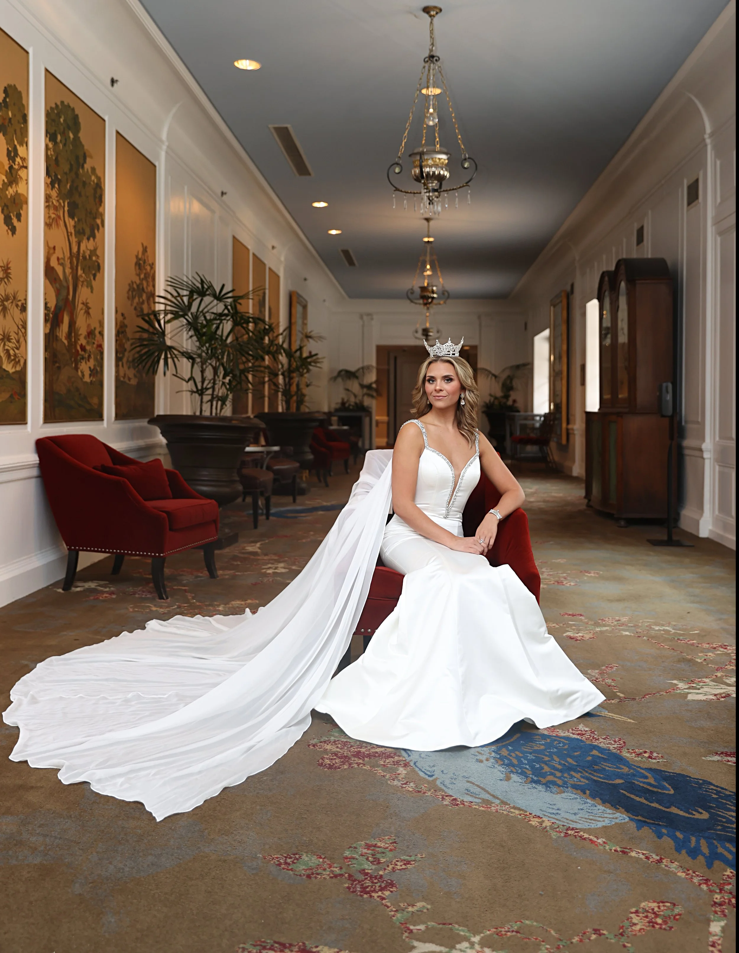 A woman in a wedding dress wearing a crown, sitting on a red velvet sofa in an elegant hallway with paintings, plants, and chandeliers.