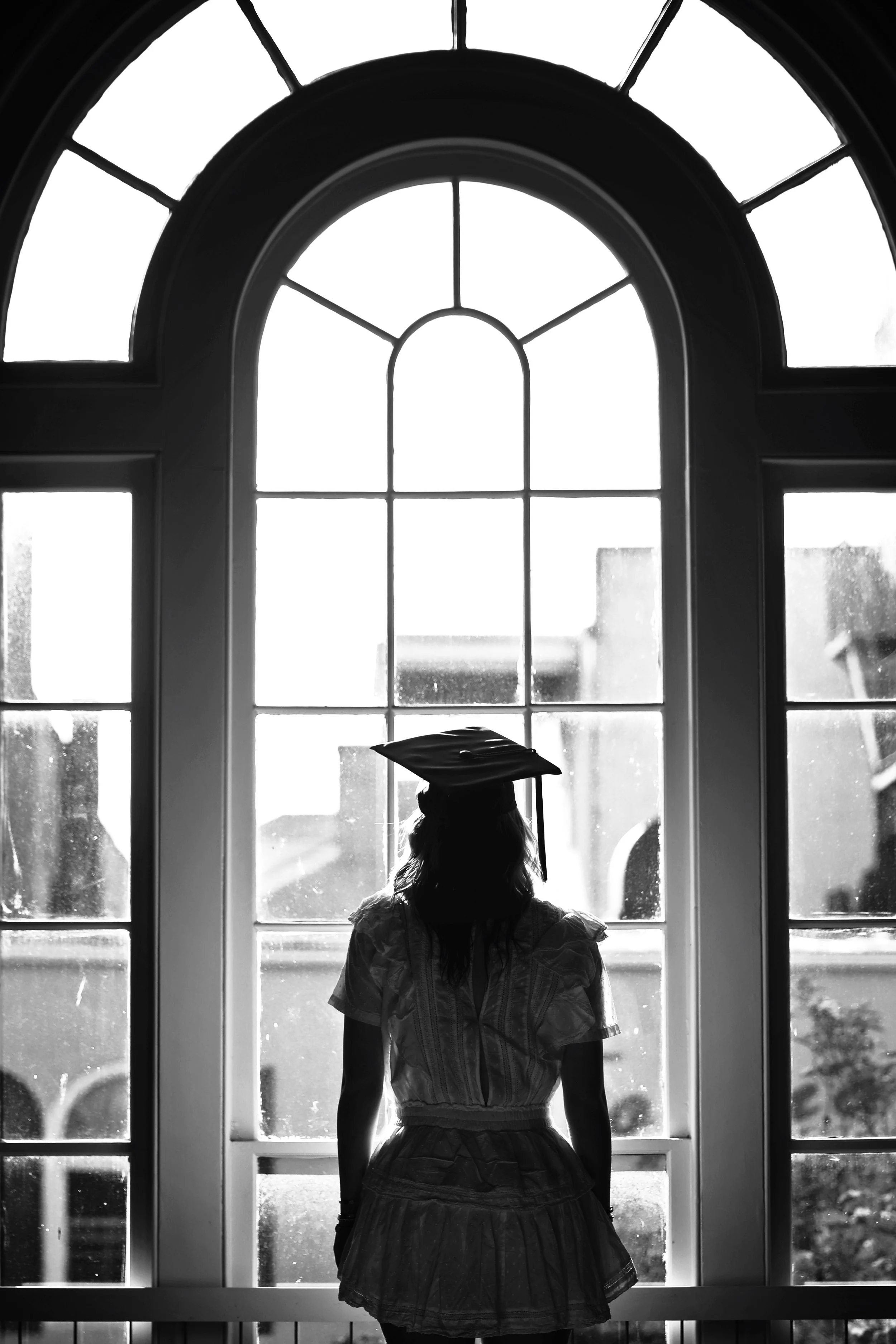 A person wearing a graduation cap looking out of a large arched window.