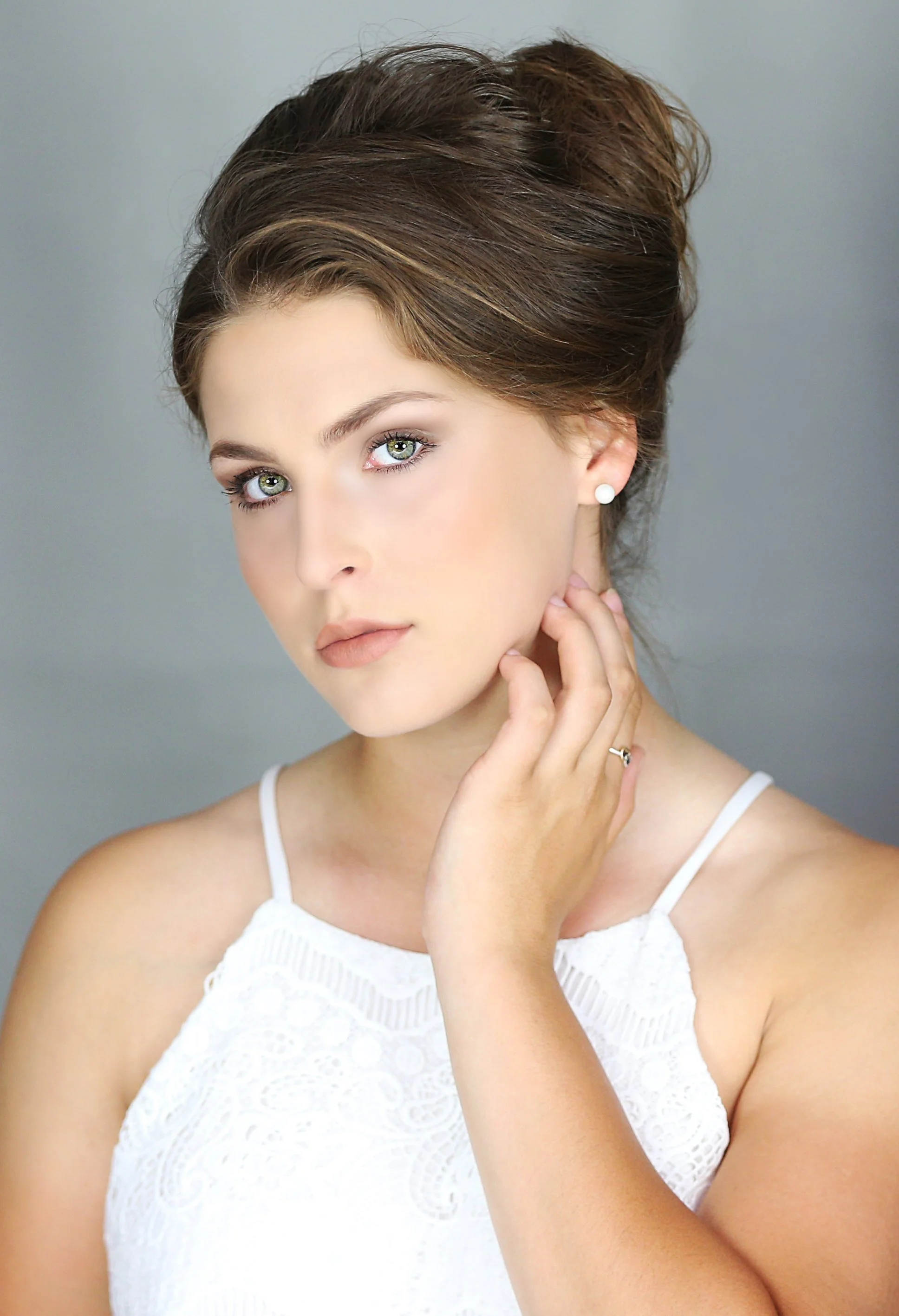 A woman with styled brown hair, blue eyes, and makeup, wearing a white lace sleeveless top and pearl earrings, posing with her hand touching her neck, against a gray background.