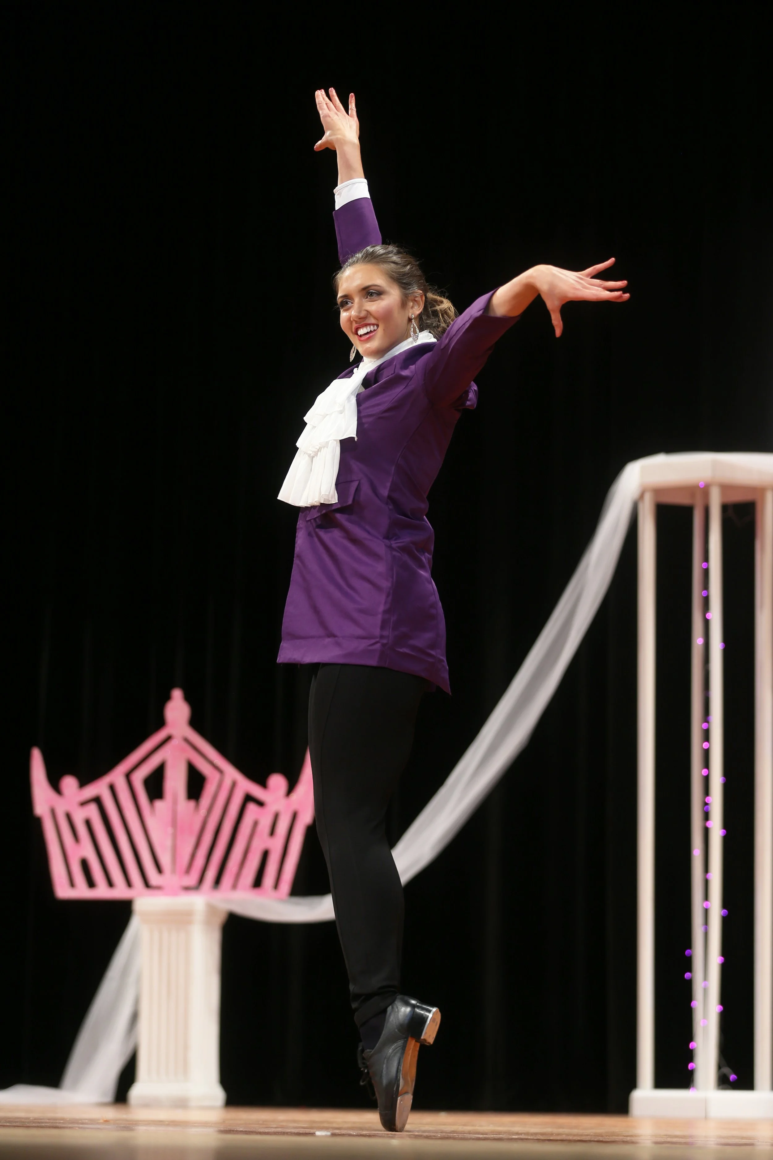 A woman performing a dance move on stage with arms raised, wearing a purple jacket, black pants, and black shoes, with stage decorations including a pink crown and white drapes.