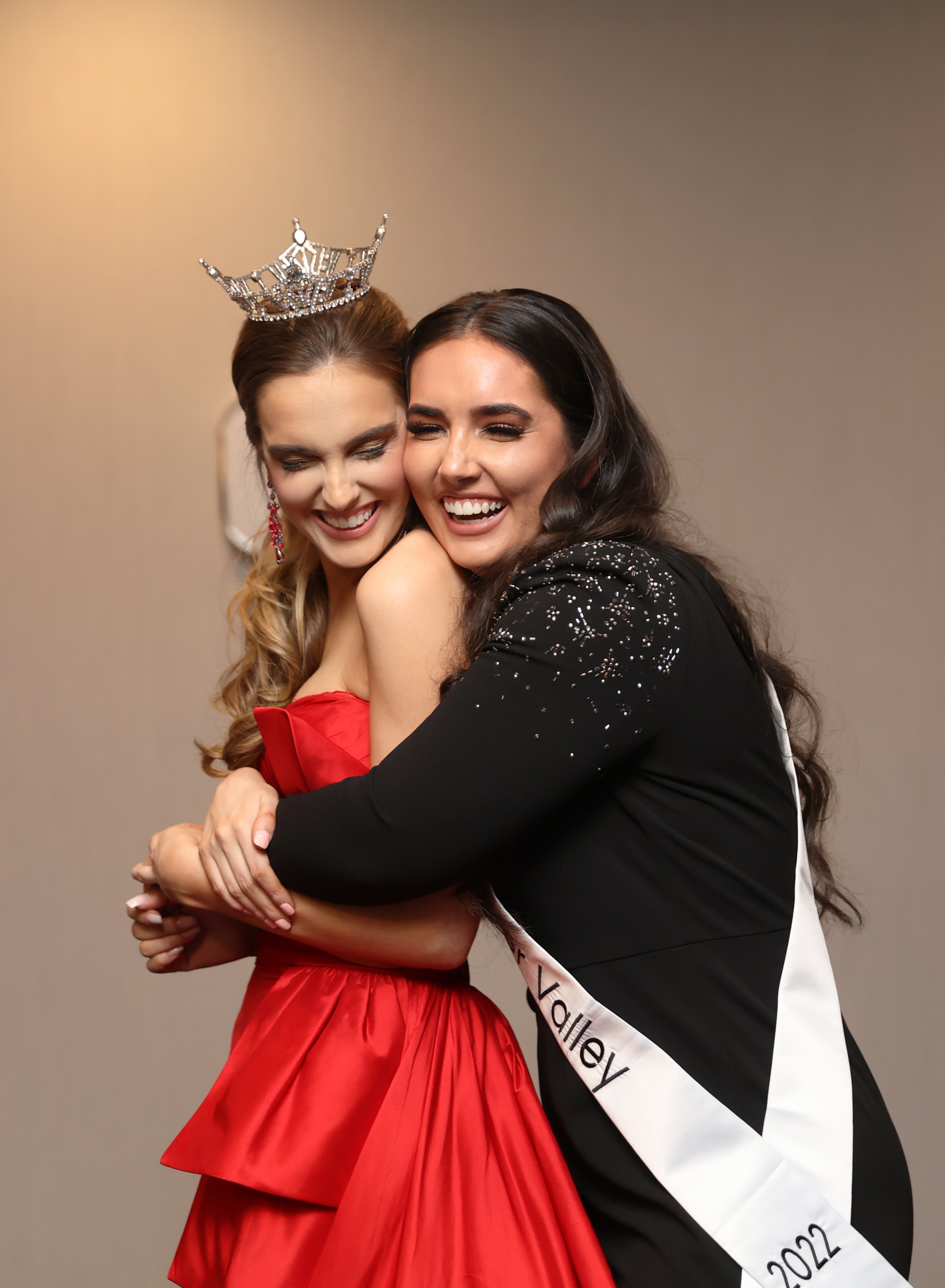 Two women hugging and smiling, one wearing a sash that says 'Valley 2022' and a crown, and the other wearing a black dress with sequins.