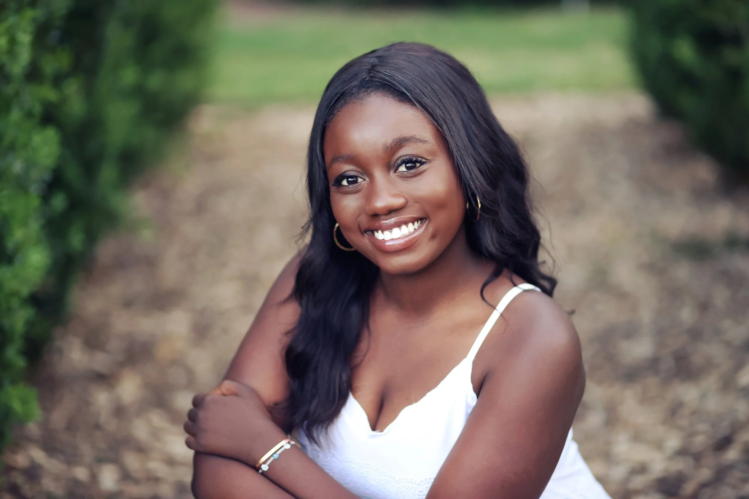 A young woman with long dark hair, wearing a white sleeveless top, smiling outdoors with green bushes in the background.