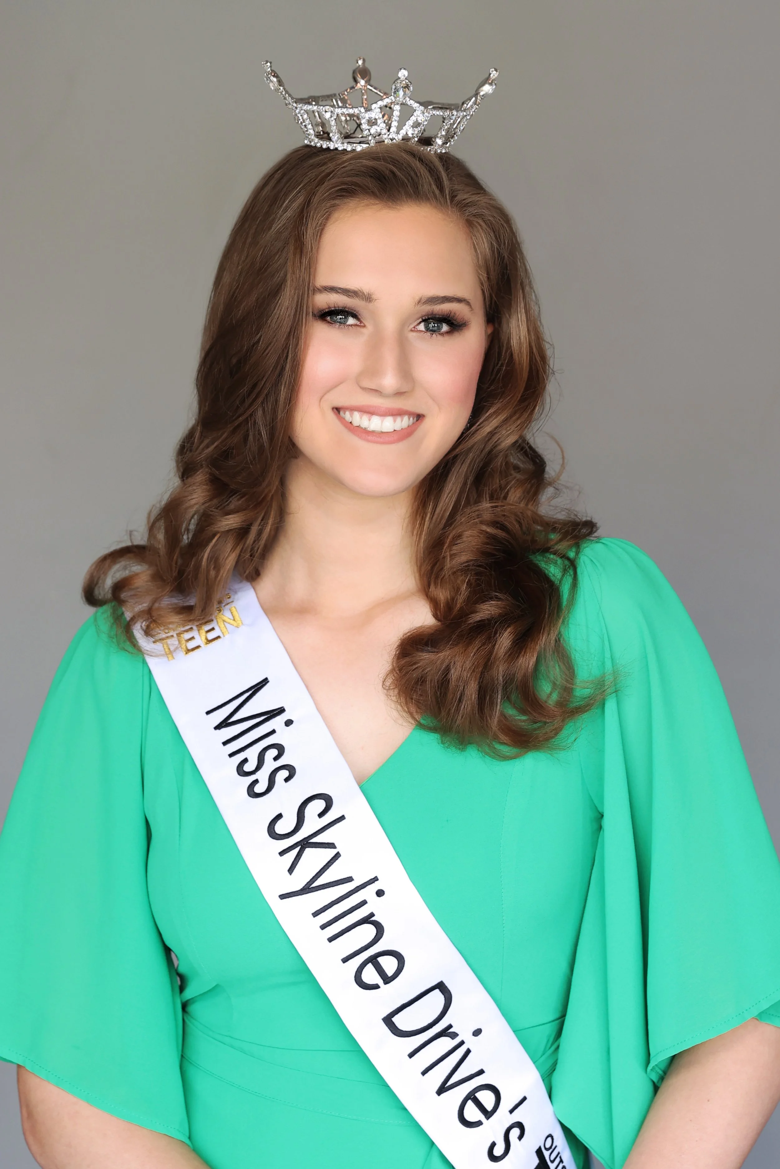 Young woman with brown, curly hair wearing a green dress, a crown, and a sash that reads 'Miss Skyline Drives' and 'Teen'.