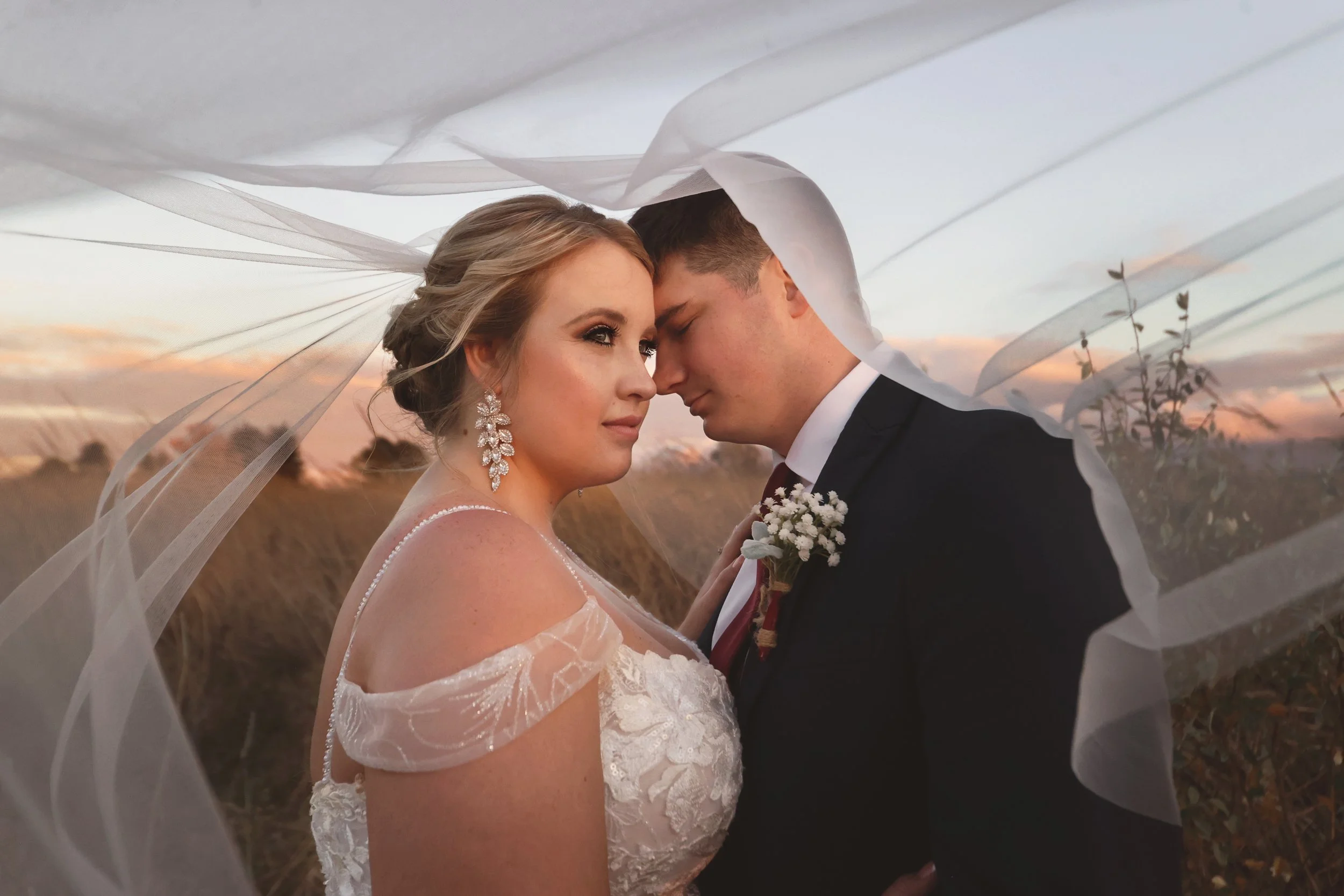 A bride and groom stand close together under a flowing veil during sunset in a field, with the bride wearing a lace wedding dress and large earrings, and the groom in a suit with a boutonniere.