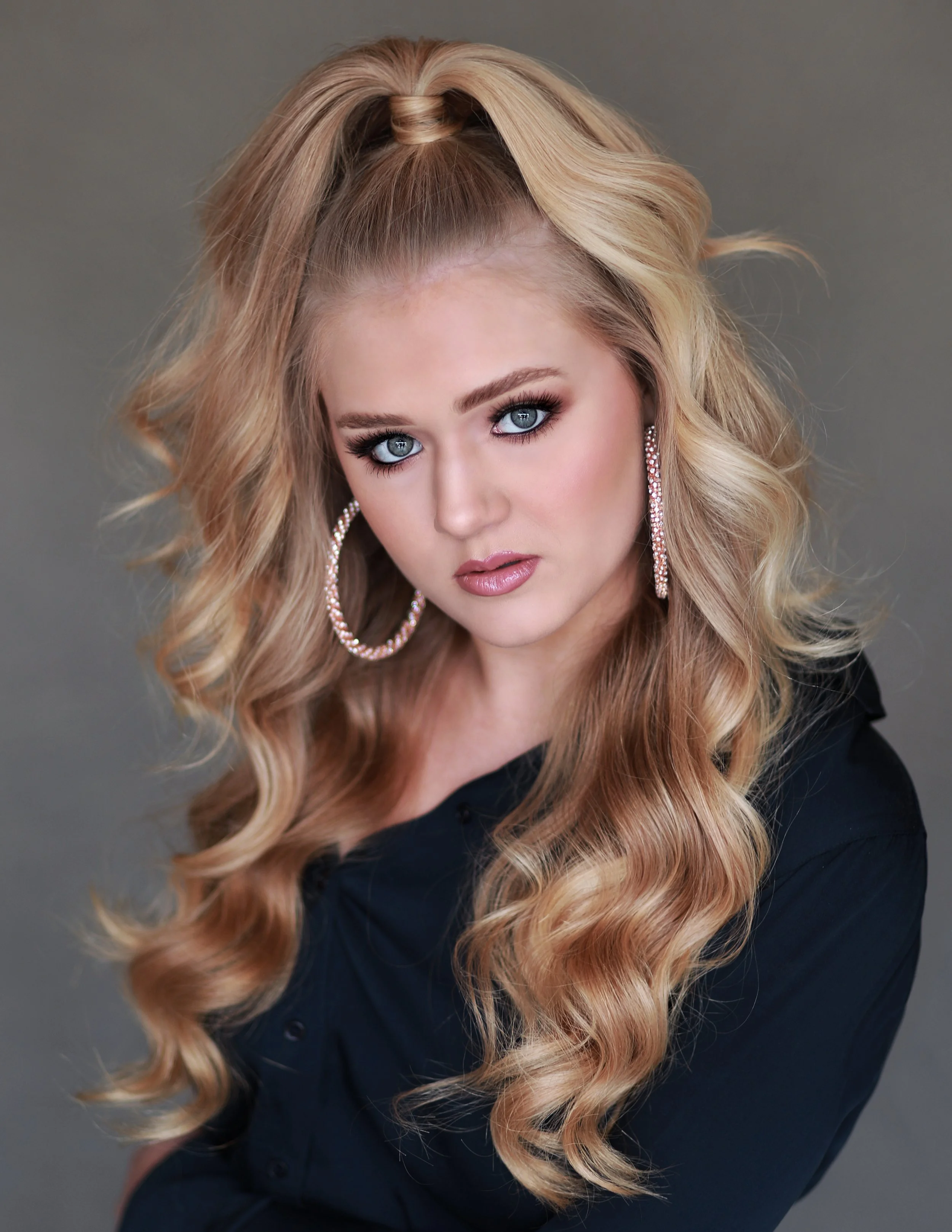 A woman with long, wavy blonde hair styled with a high ponytail, wearing large hoop earrings and makeup, dressed in a black top, looking directly into the camera against a gray background.