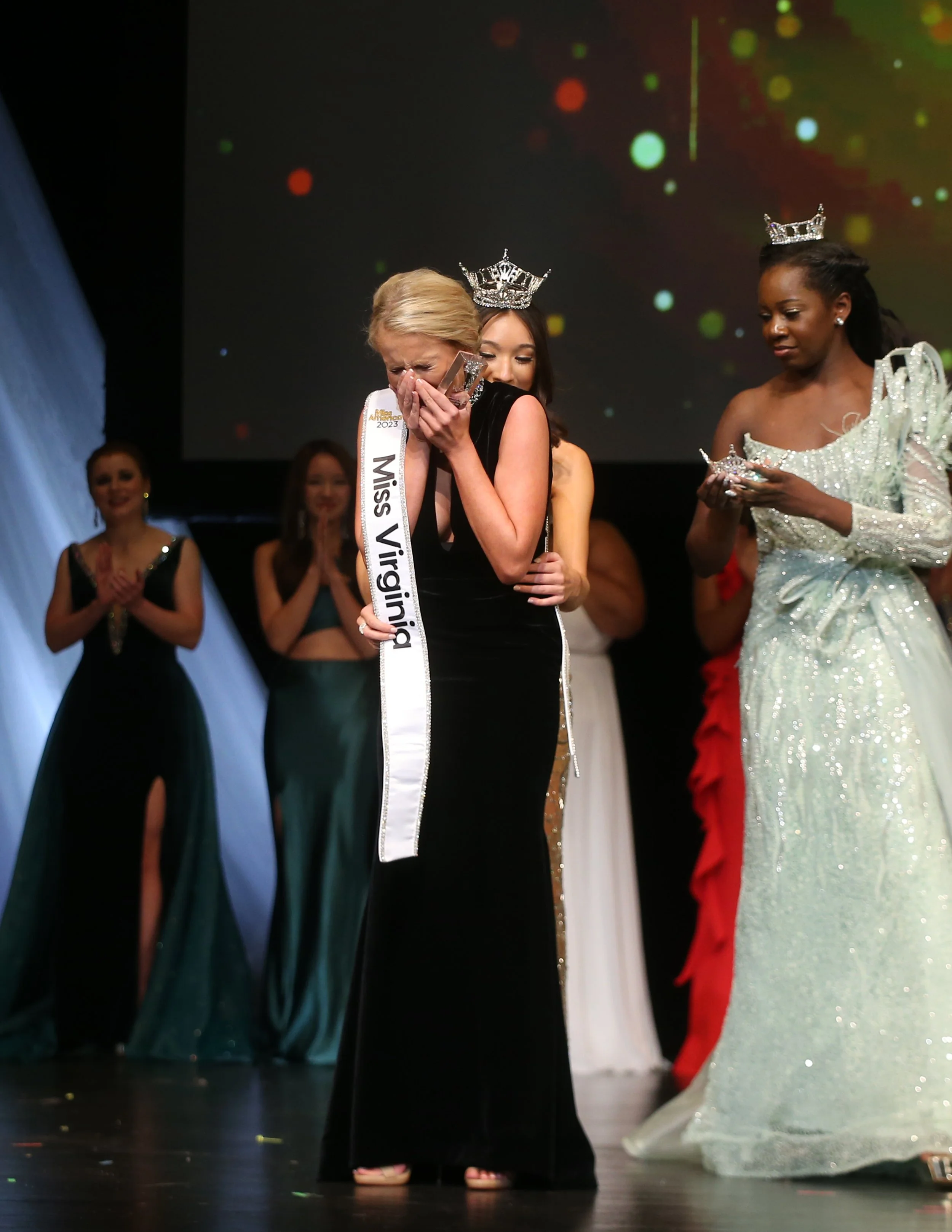 Miss Virginia wearing a crown and sash, visibly emotional, being crowned as winner during a beauty pageant on stage with other contestants in evening gowns.