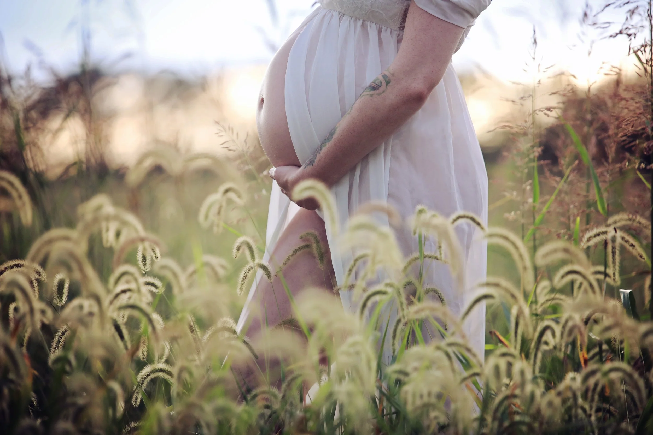 Pregnant woman in a white dress standing in a field of tall grass during sunset, holding her belly with both hands.