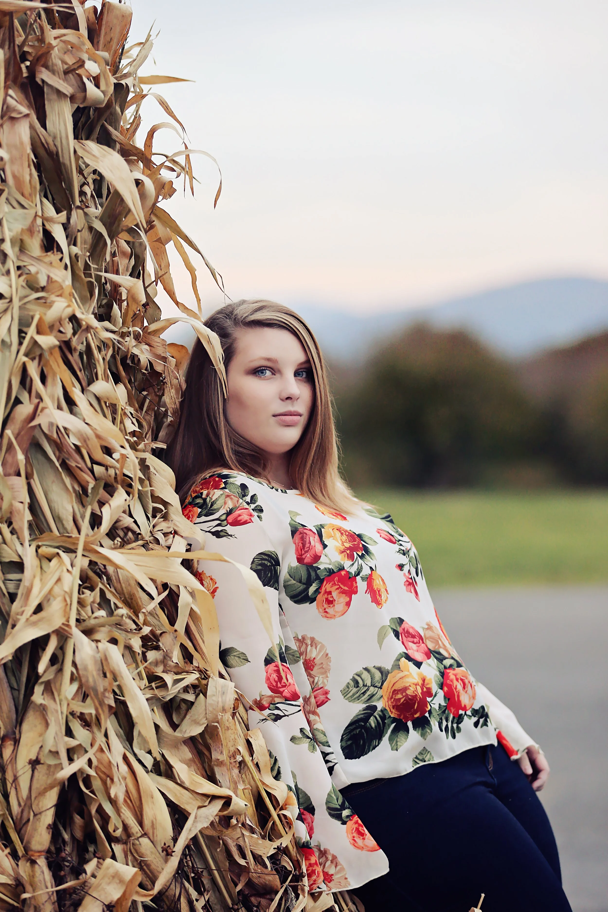 A young woman with blonde hair and blue eyes leaning against a haystack outdoors, wearing a white blouse with a red and green floral pattern and dark pants.