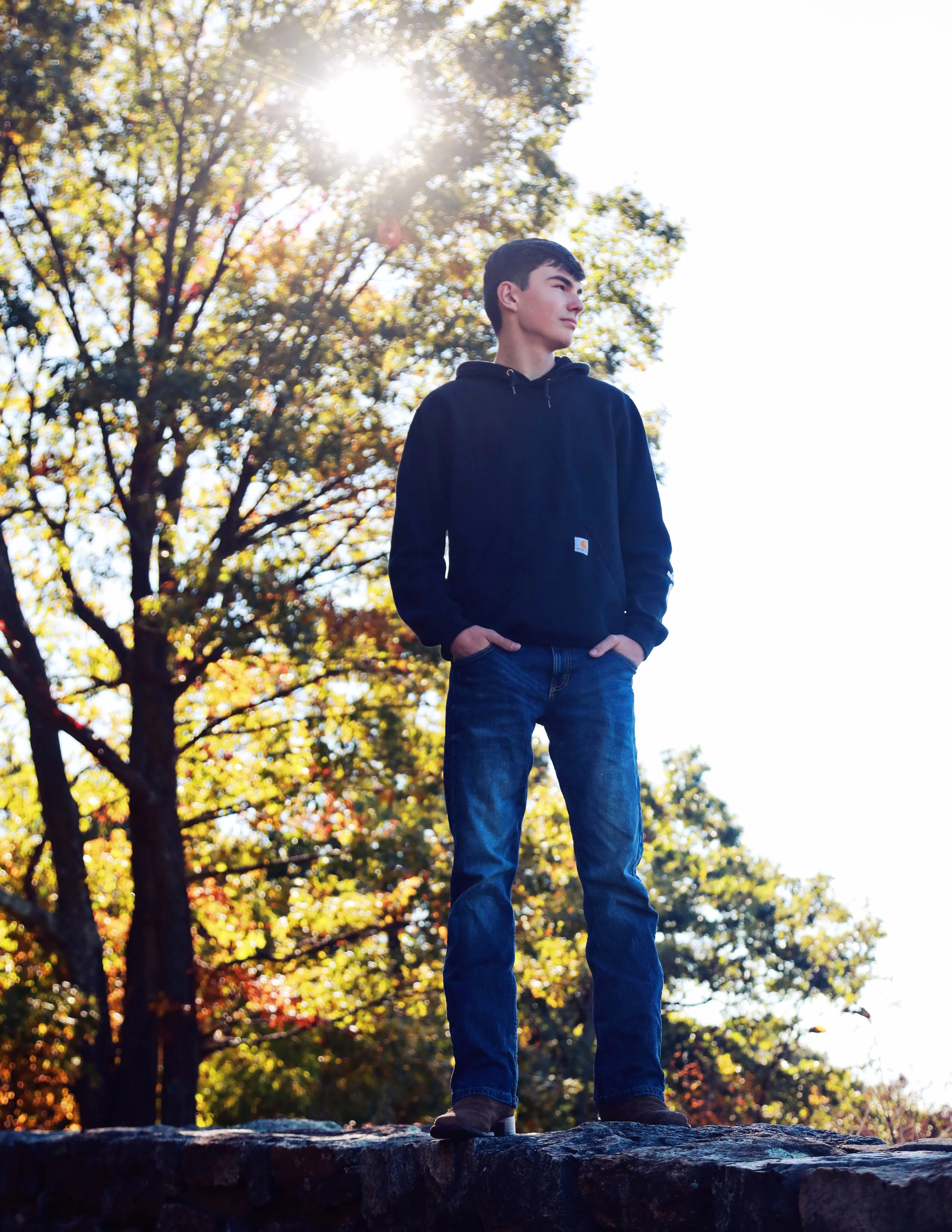 A young man standing outdoors on a rocks, wearing a black hooded sweatshirt and jeans, with his hands in his pockets, looking to the right. In the background, there are trees with autumn leaves and the sun shining through the branches.