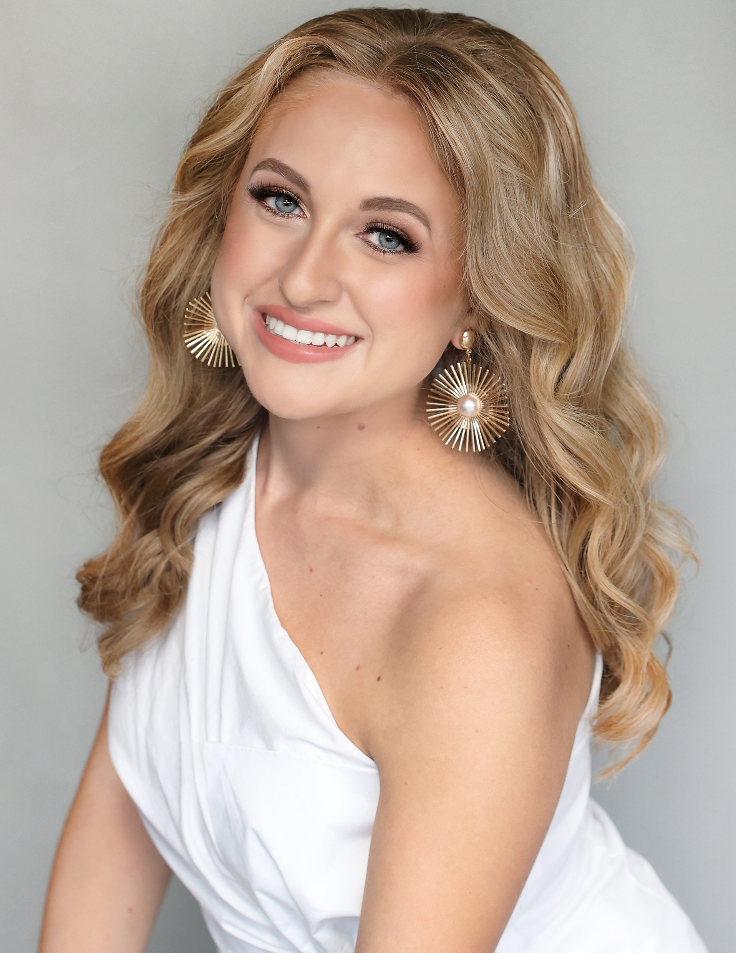 A woman with blonde, wavy hair, blue eyes, and makeup, smiling and wearing white clothing and large gold disc-shaped earrings.