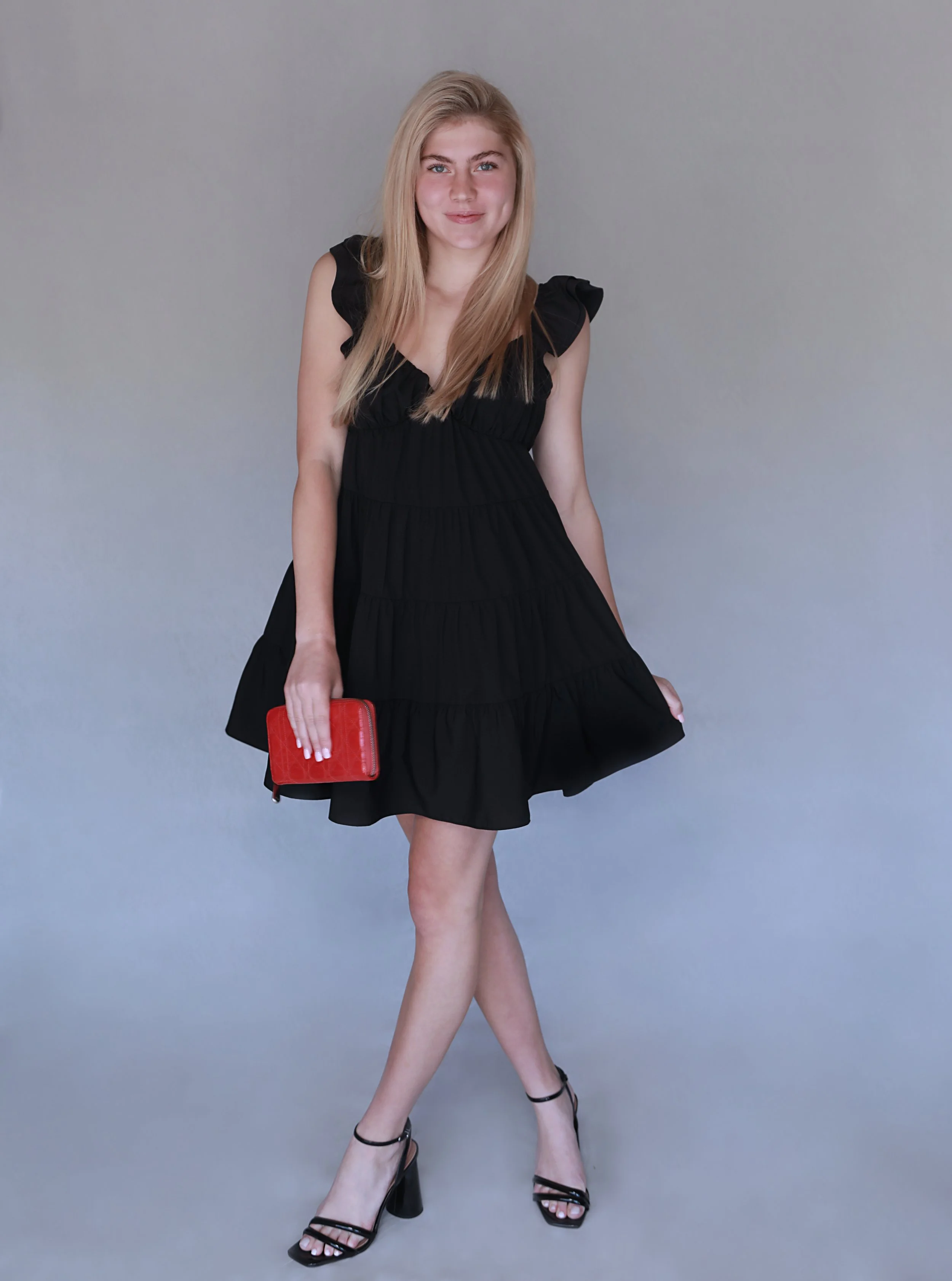 A young woman in a black dress holding a red clutch purse and wearing black high-heeled sandals, standing against a plain gray background.