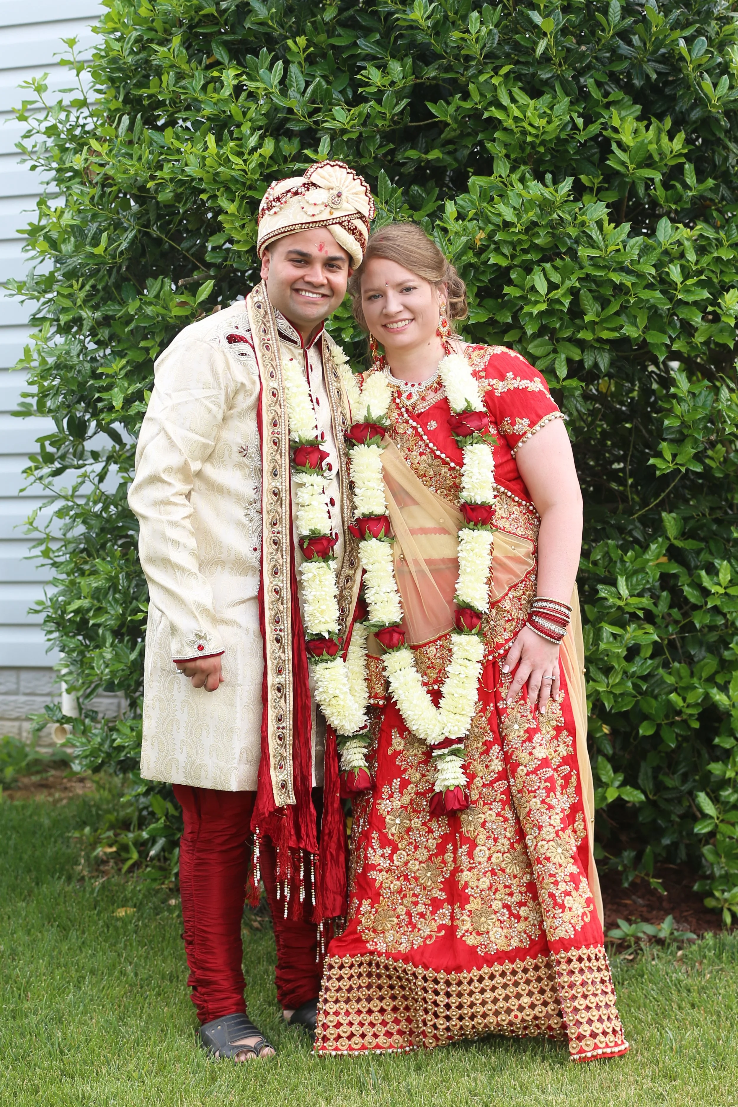 A newlywed couple in traditional Indian wedding attire standing outdoors in front of green bushes, smiling at the camera, with floral garlands around their necks.