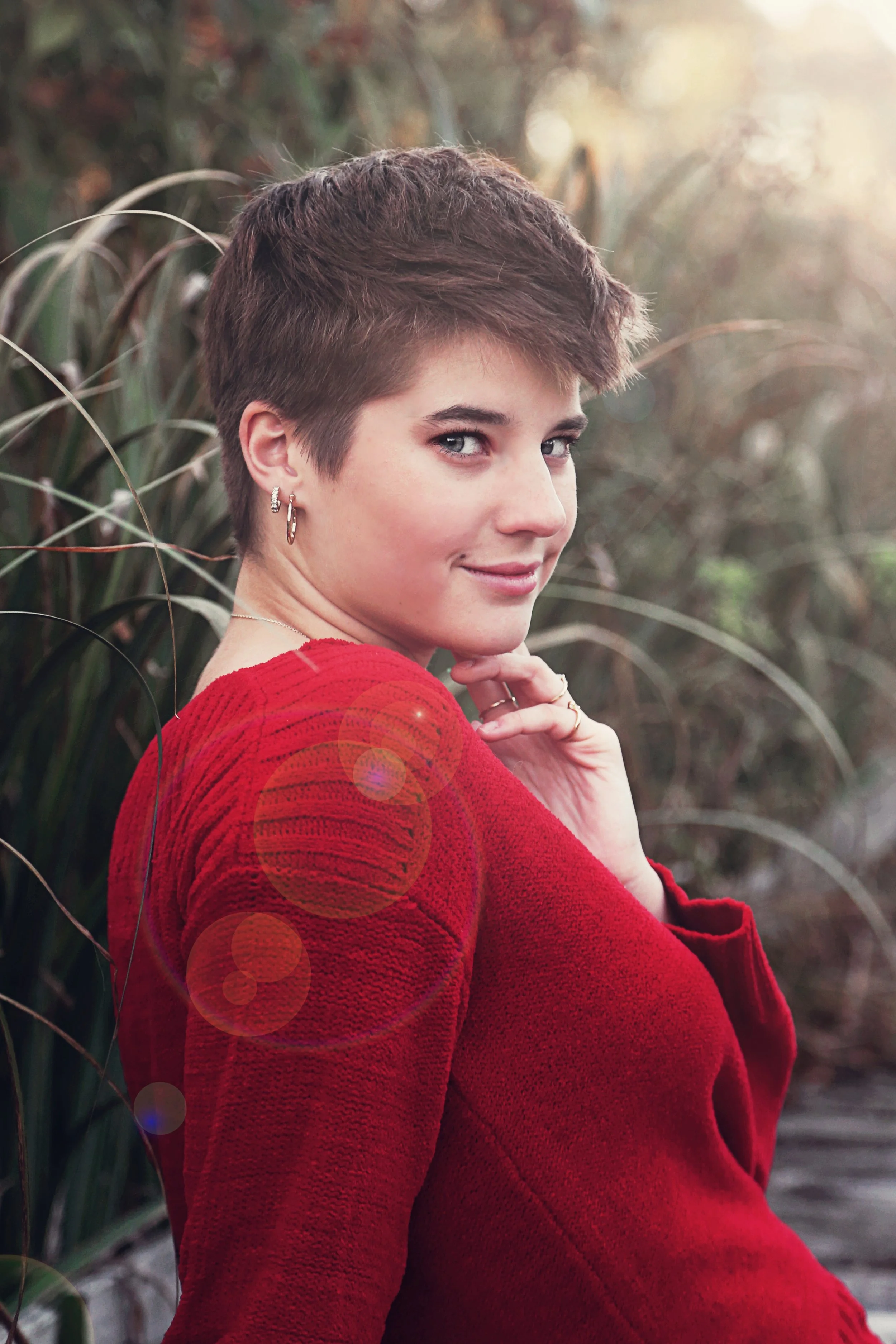 A woman with short brown hair wearing a red sweater, standing outdoors among tall grass or reeds with sunlight in the background.